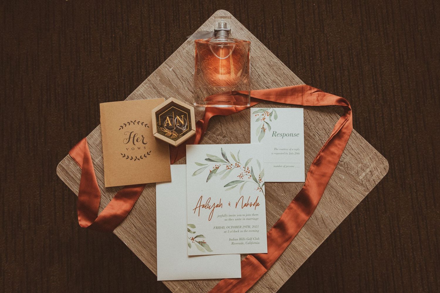 Wedding stationery featuring botanical design with perfume bottle and rust-colored ribbon on wooden board.