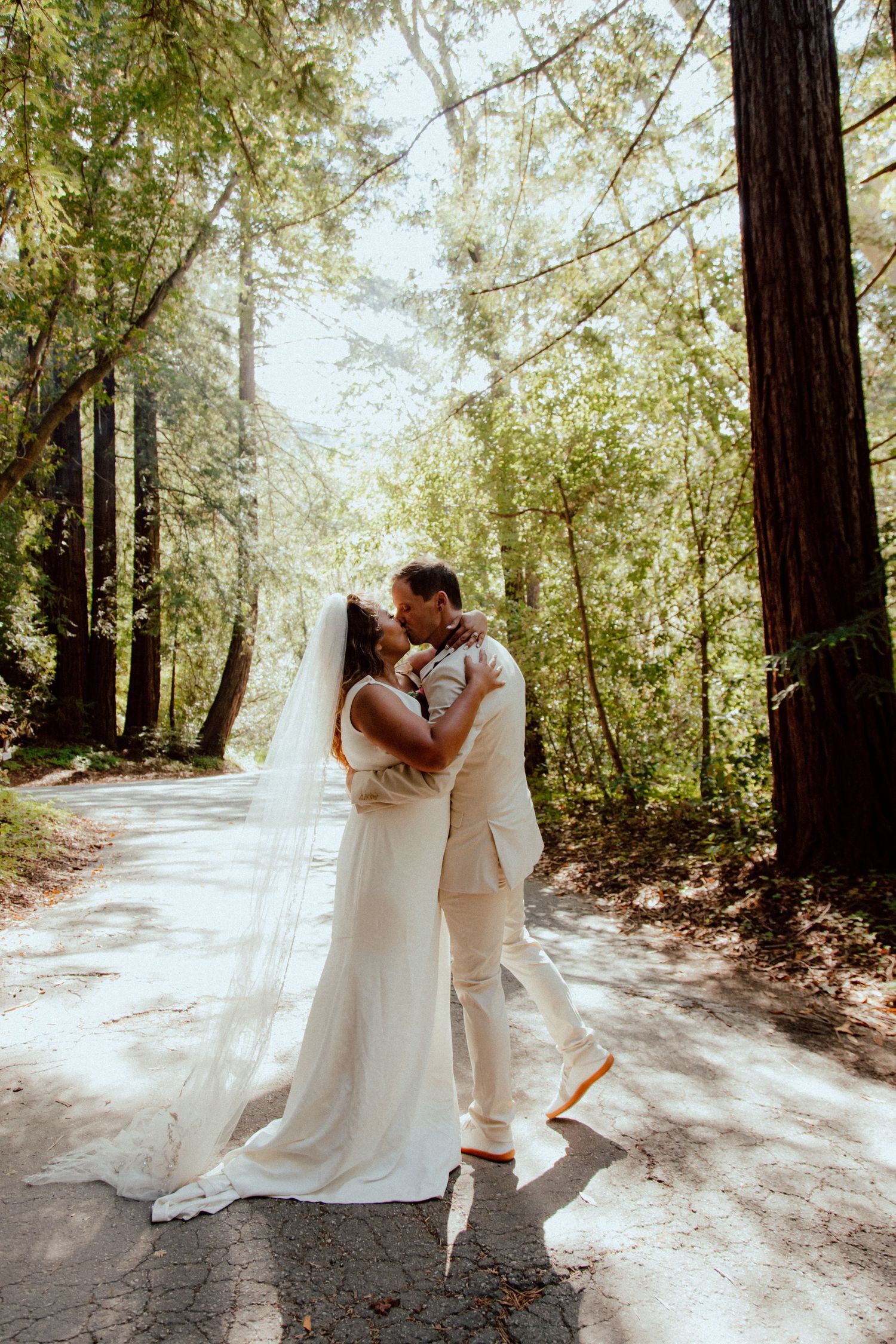 Newlyweds share an intimate moment on a sunlit forest path.