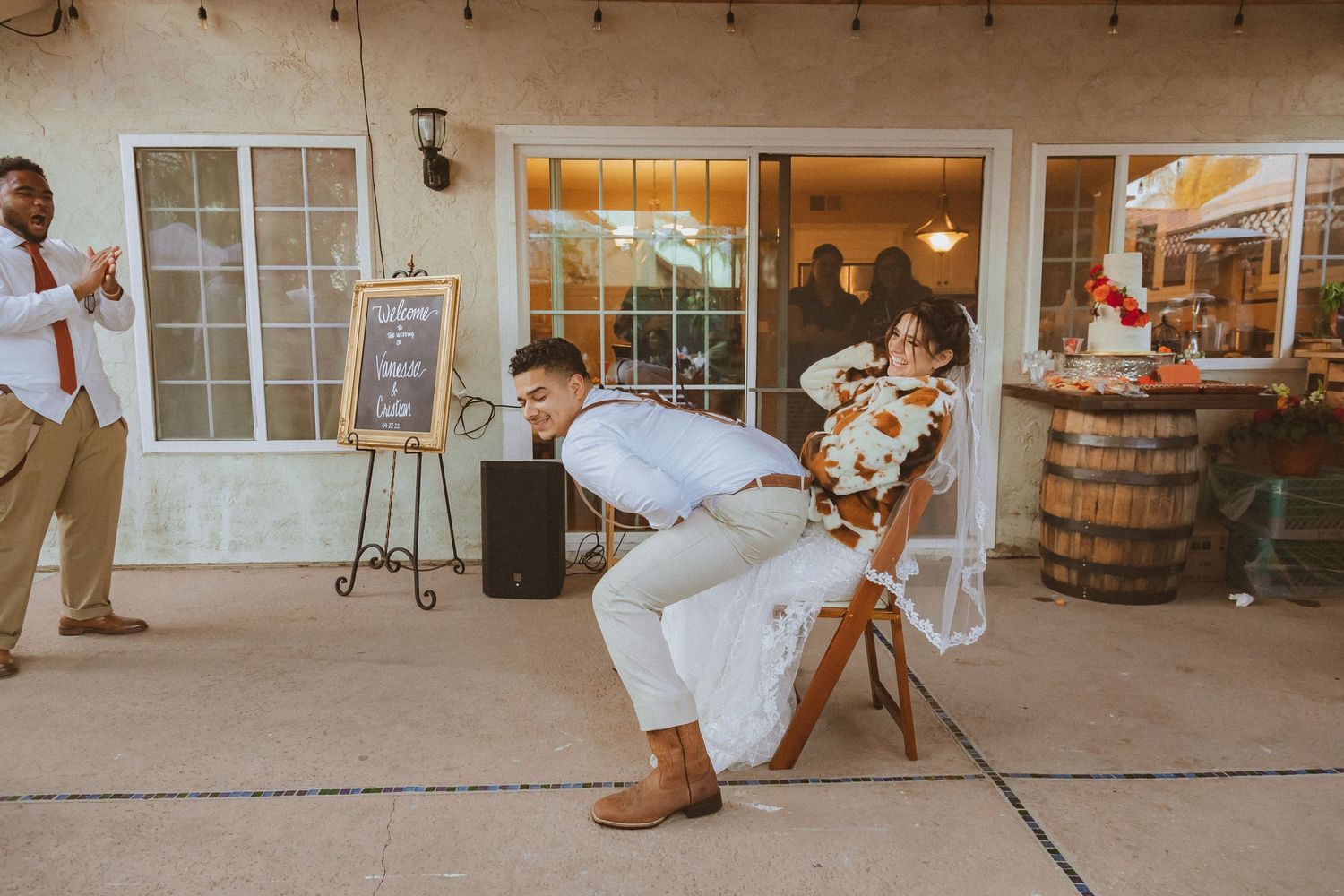 Wedding guests watch as a couple performs a playful dance move on a covered outdoor patio.