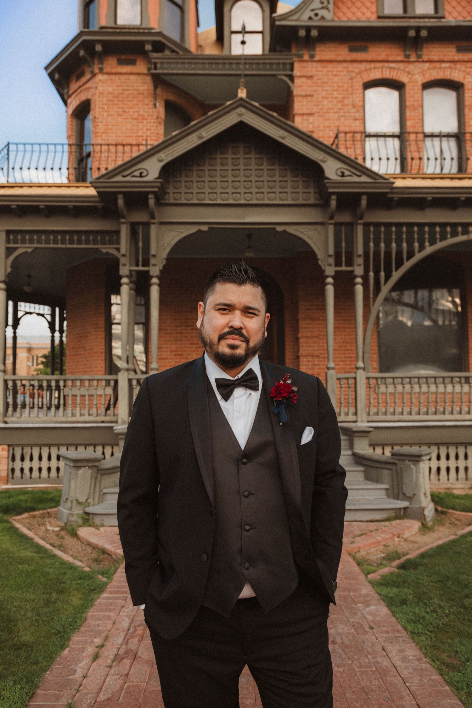 A formal portrait in front of a historic red brick Victorian mansion with ornate architectural details.