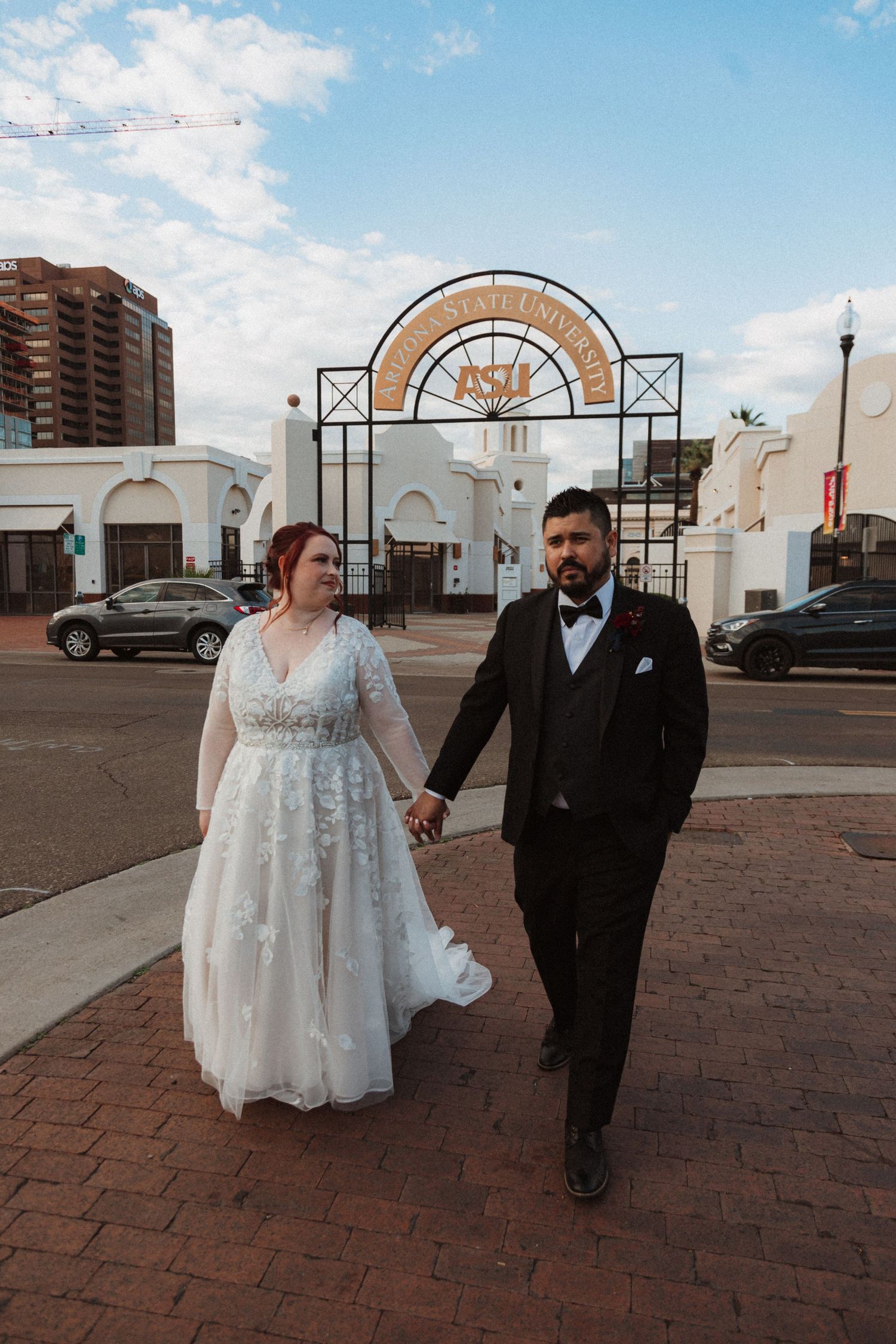 Wedding couple strolls together on brick walkway with decorative metal arch and urban backdrop.
