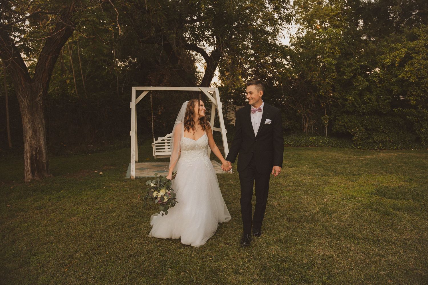 Wedding couple poses in front of a simple white wooden arch on a sunny lawn surrounded by trees.