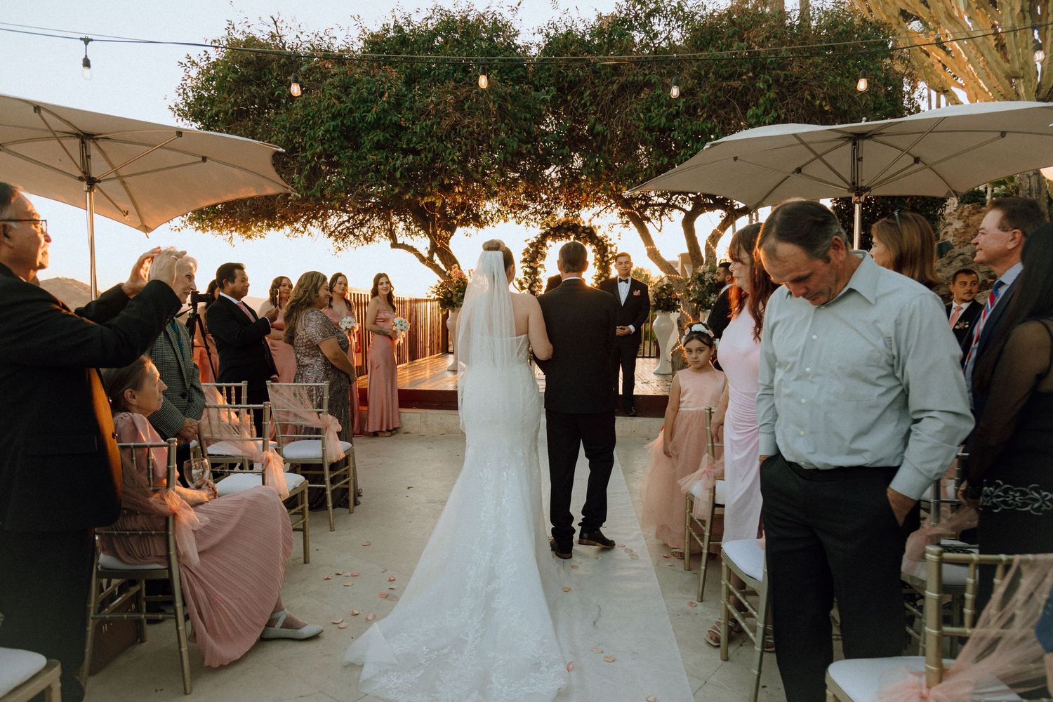 Wedding guests watch as a couple walks down the aisle at a sunset outdoor ceremony.