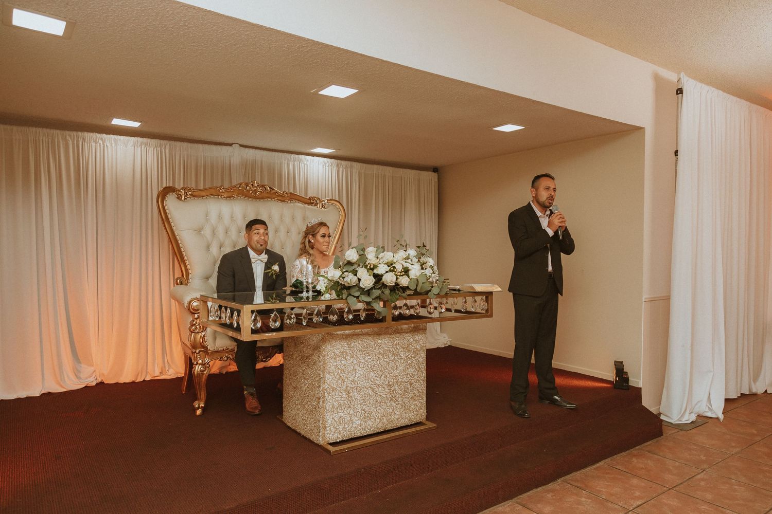 Wedding reception setup with an ornate sweetheart table and floral arrangements against white draping.
