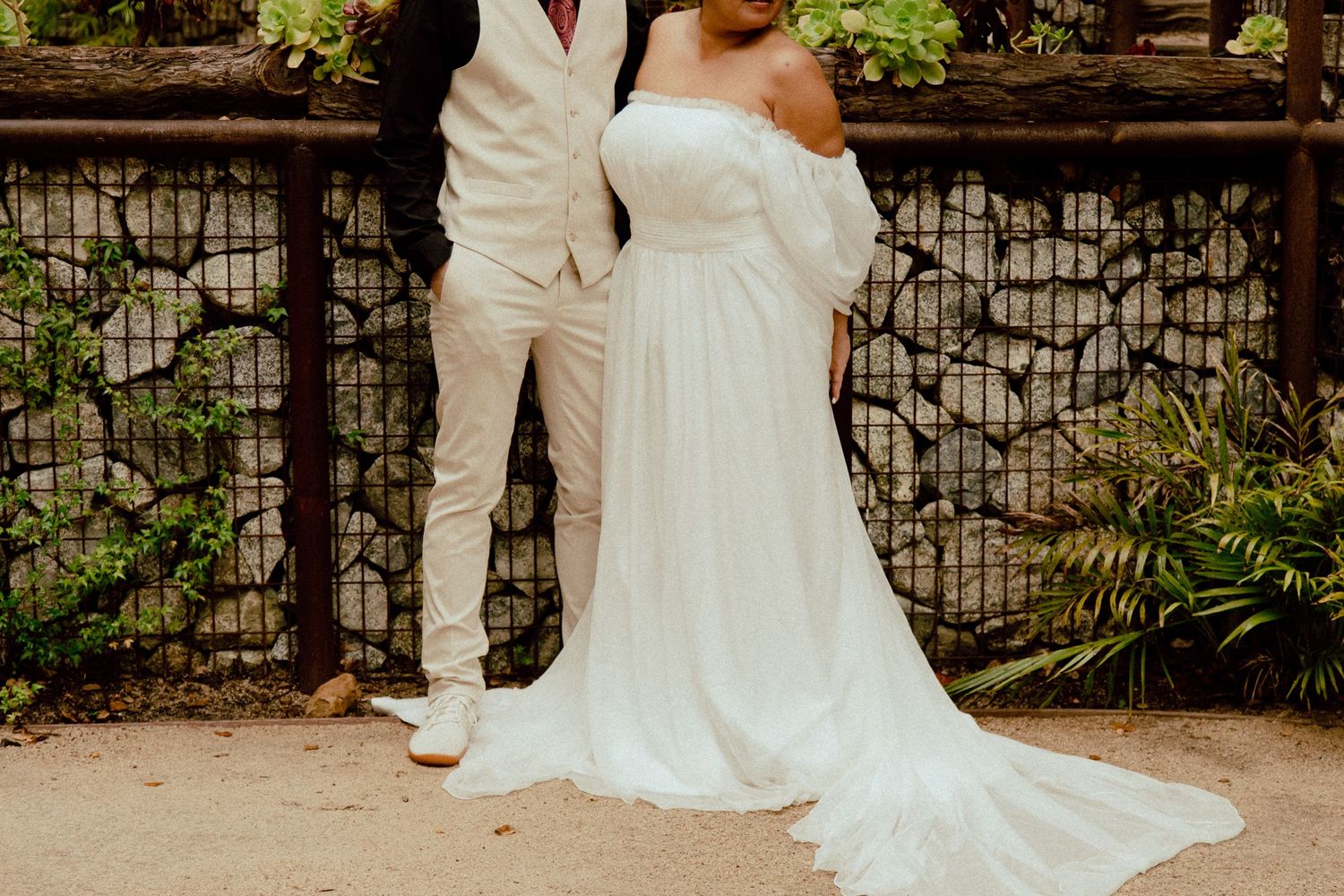 Wedding couple poses against metal and stone fence showcasing the bride's flowing white dress train.