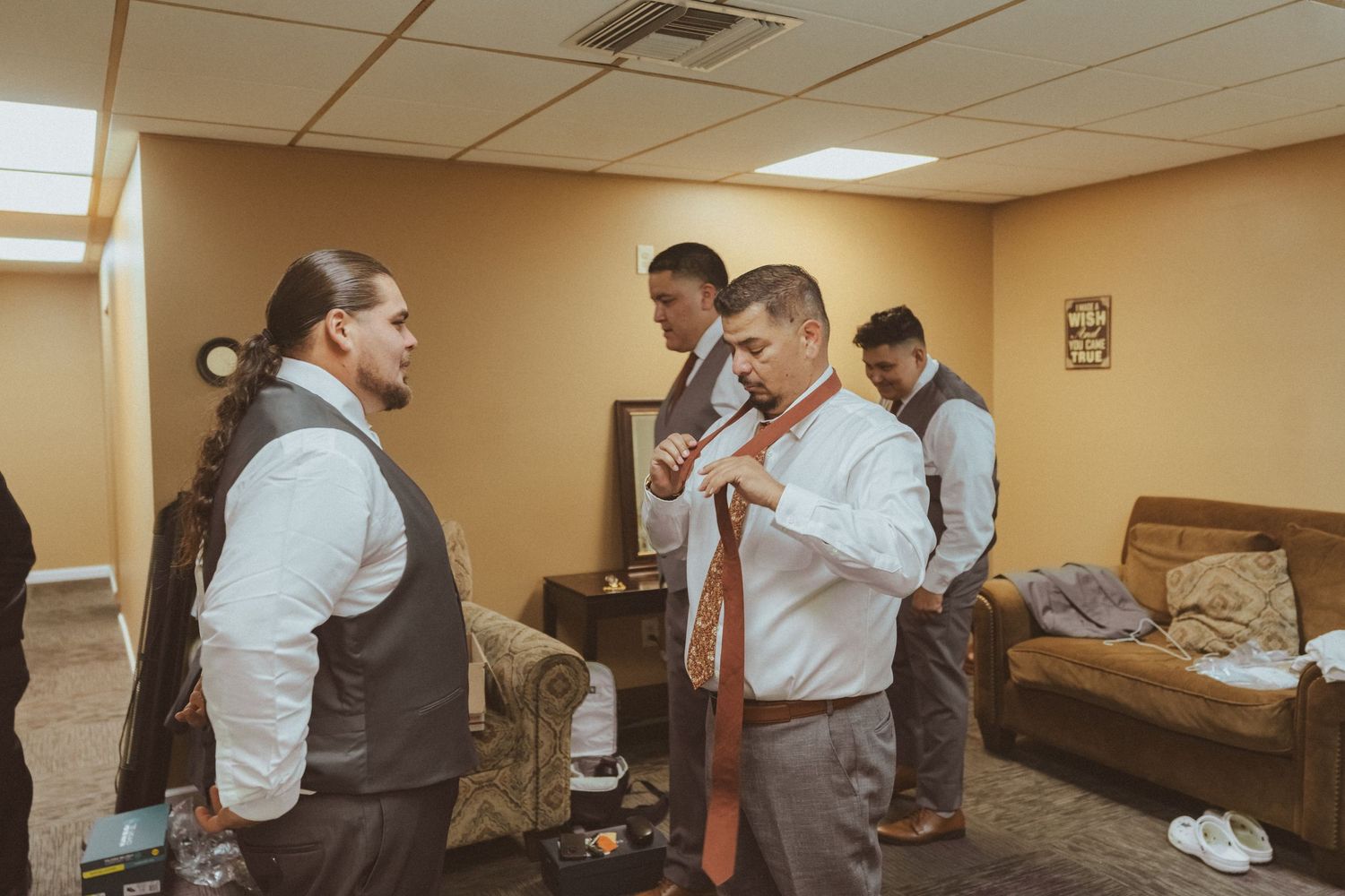 Group of formally dressed people gather in preparation room with beige walls and overhead lighting.