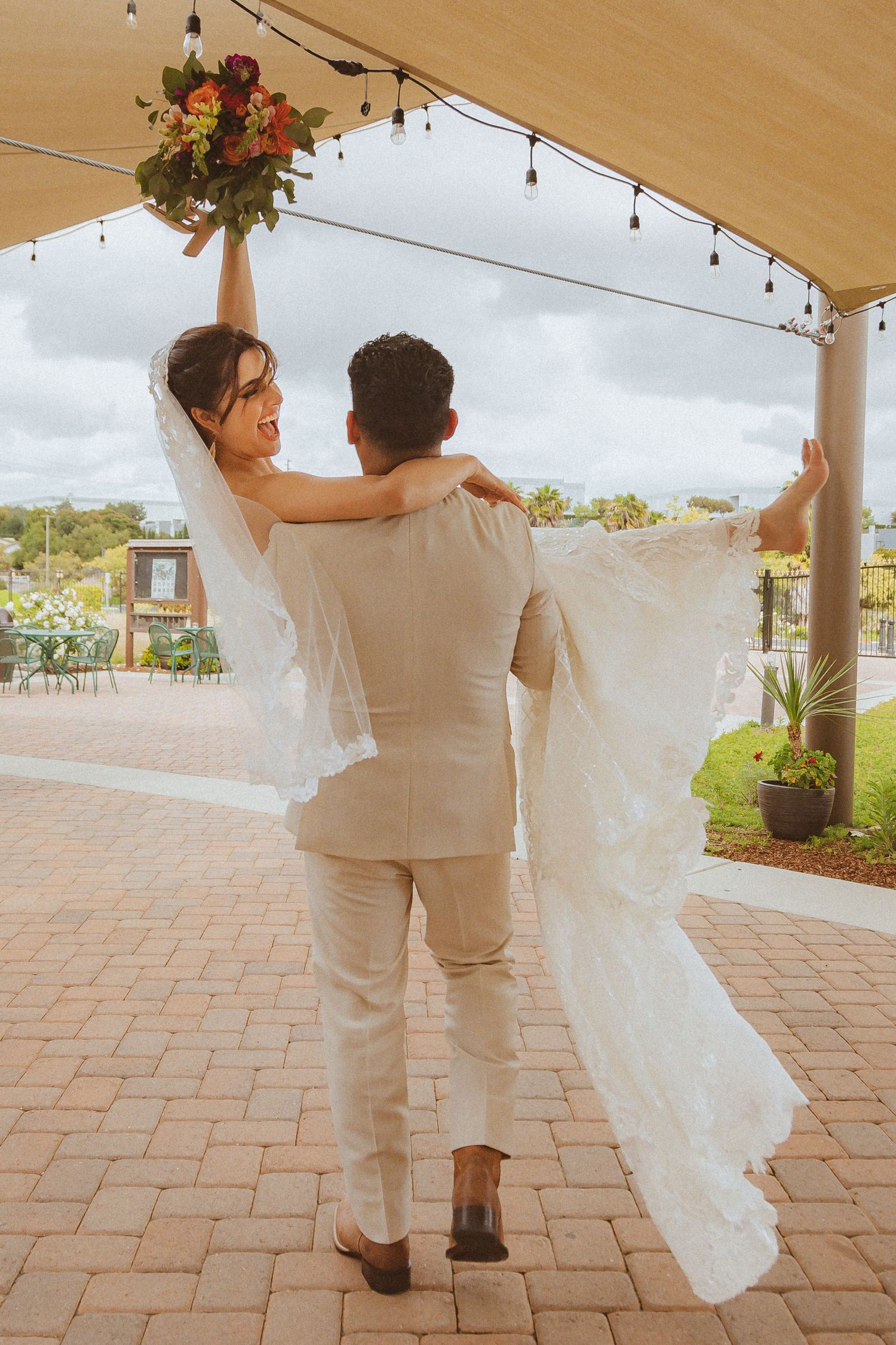 Wedding couple celebrates with raised bouquet while dancing outdoors under covered patio with string lights.