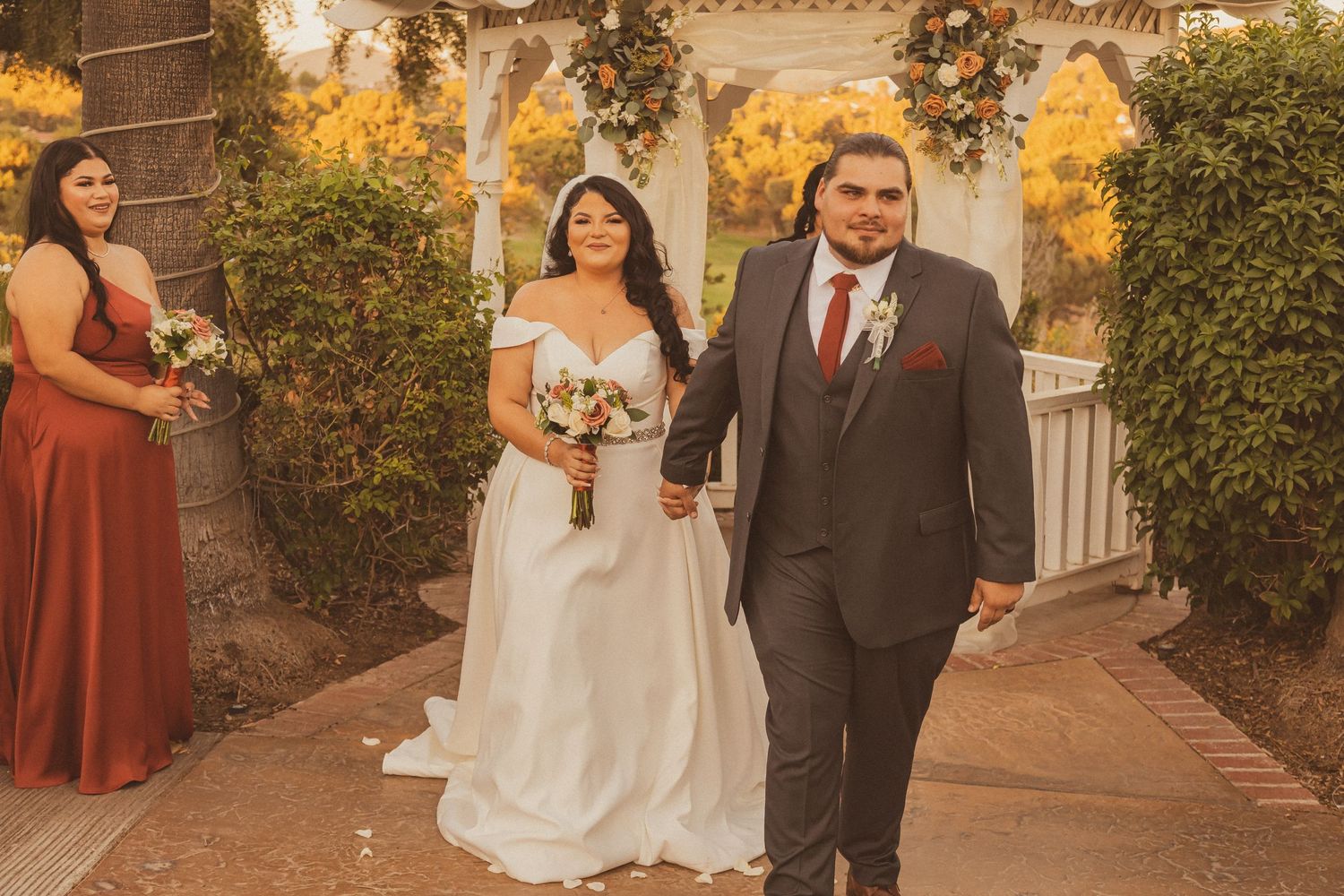 A wedding procession walks past a decorative gazebo with warm sunset lighting.