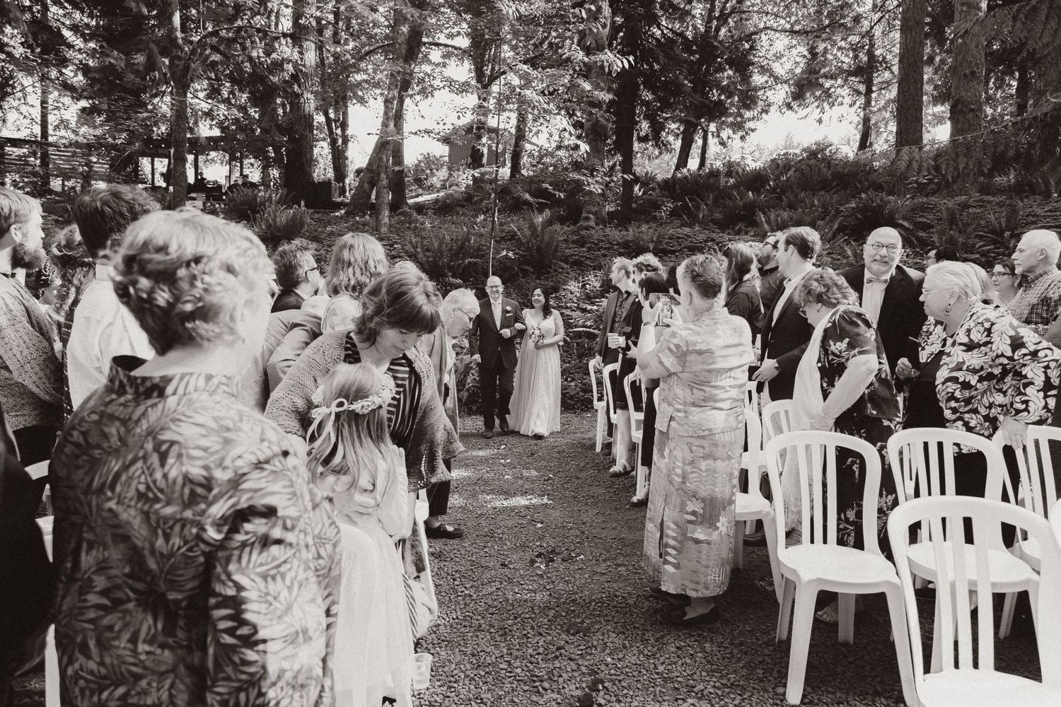 Black and white photo of wedding guests gathered on white chairs in a forest clearing.