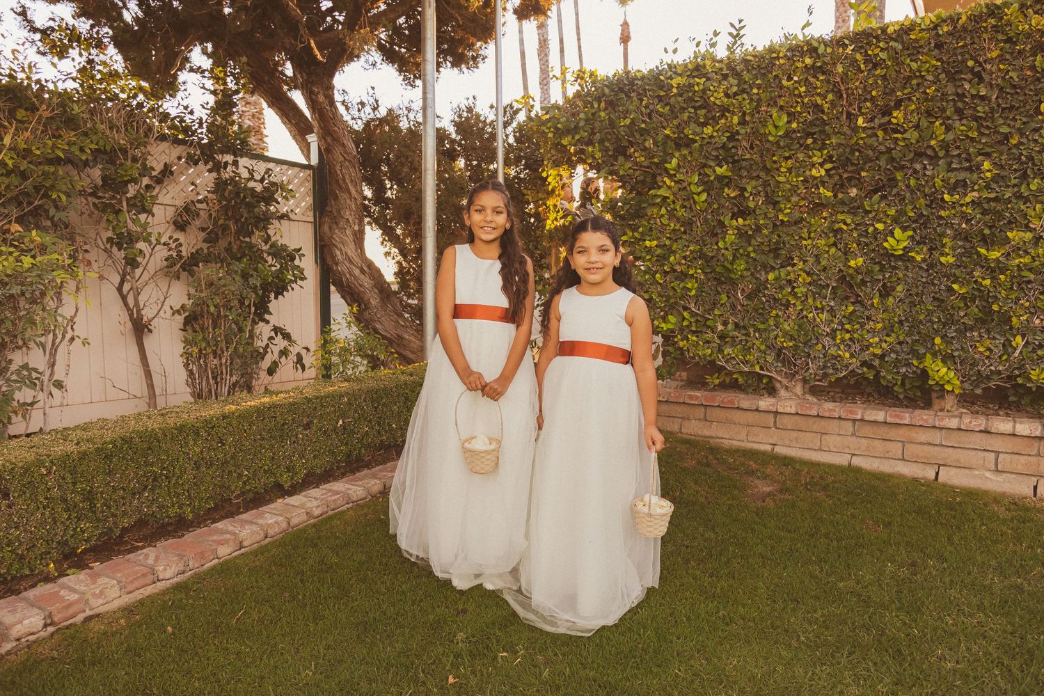 Two young attendants in matching white and orange dresses pose in a garden at sunset.