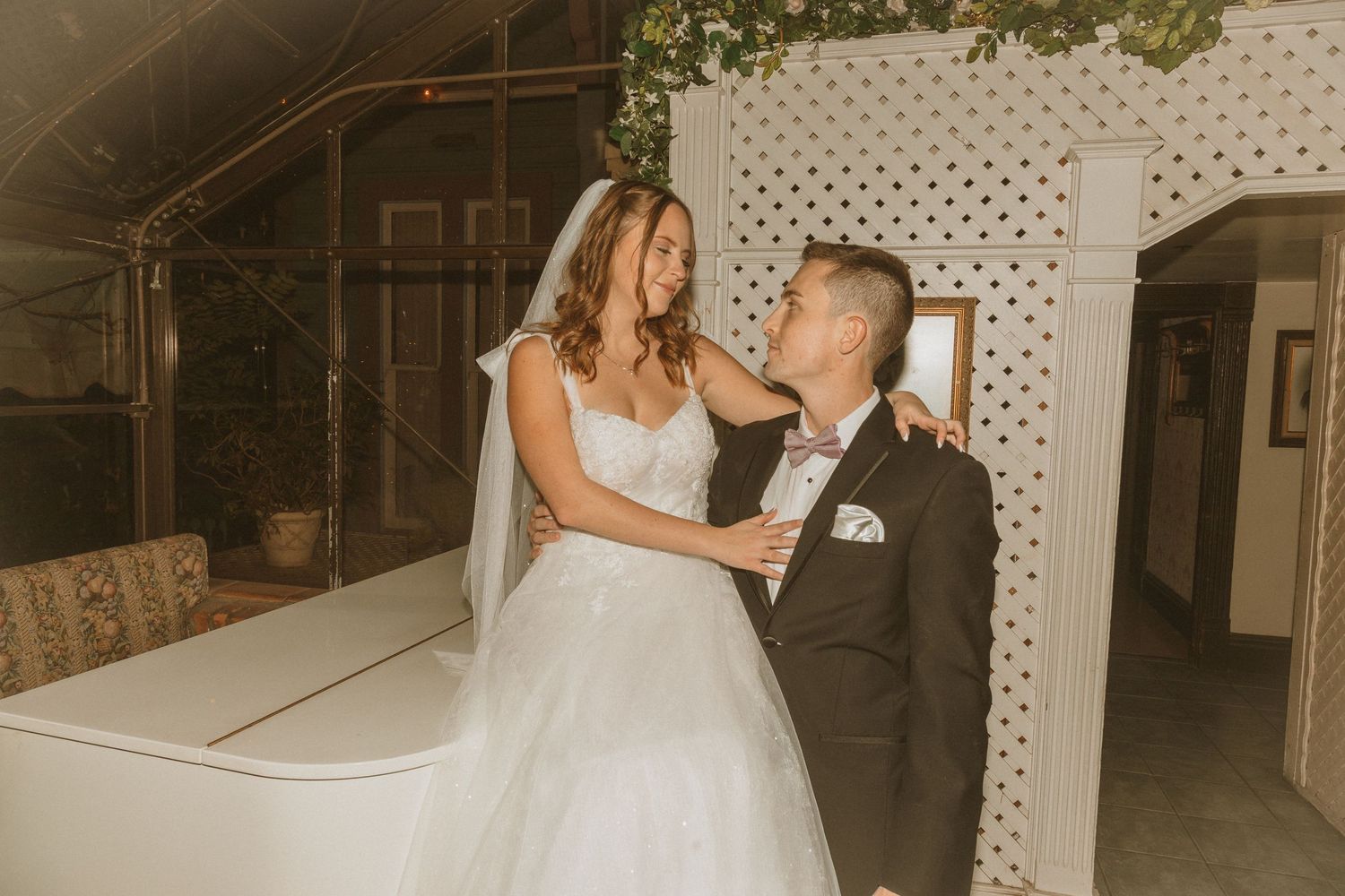 Warm-toned wedding photo of couple sharing an intimate moment beside a white piano indoors.