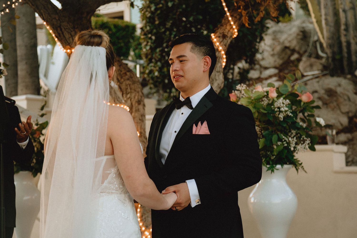 Couple shares tender moment during wedding ceremony beneath string lights and greenery.