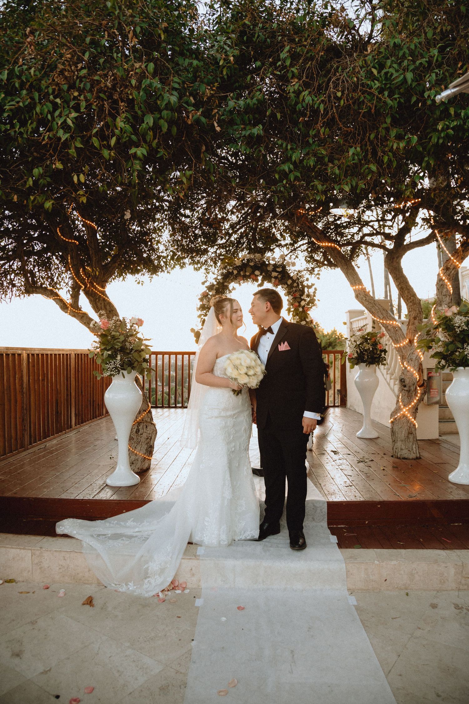 Wedding couple poses beneath overhanging trees on an outdoor terrace with elegant white decor.