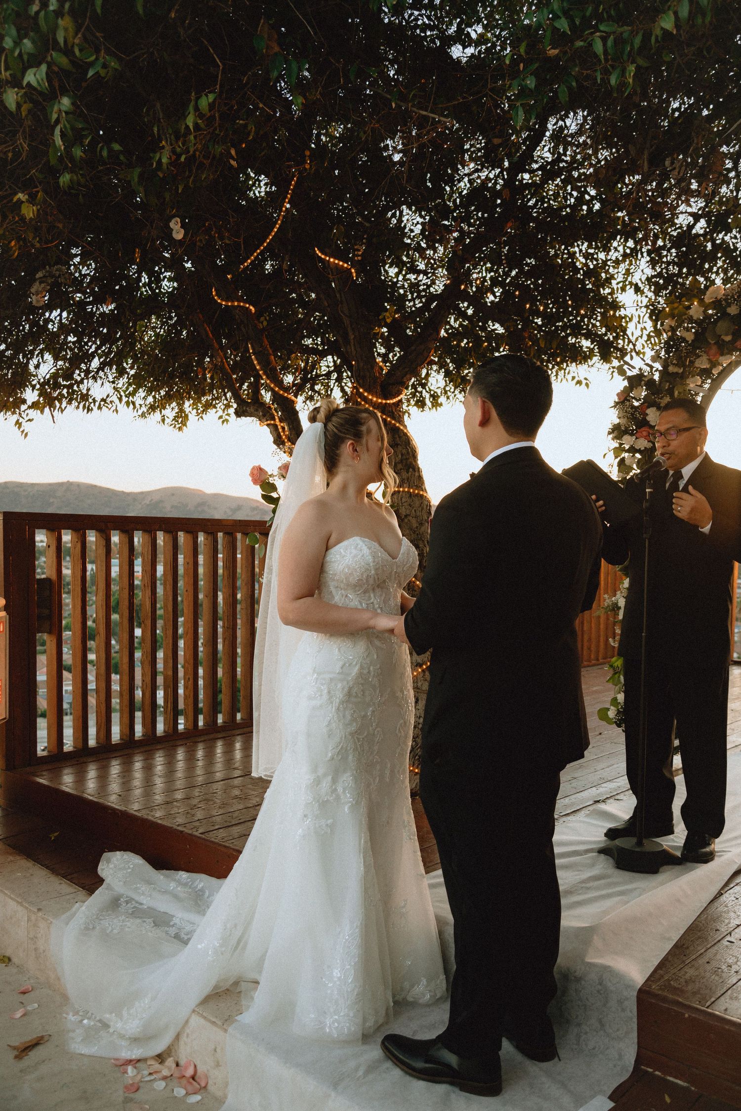 Wedding ceremony unfolds on decorated outdoor deck with mountain backdrop at sunset.
