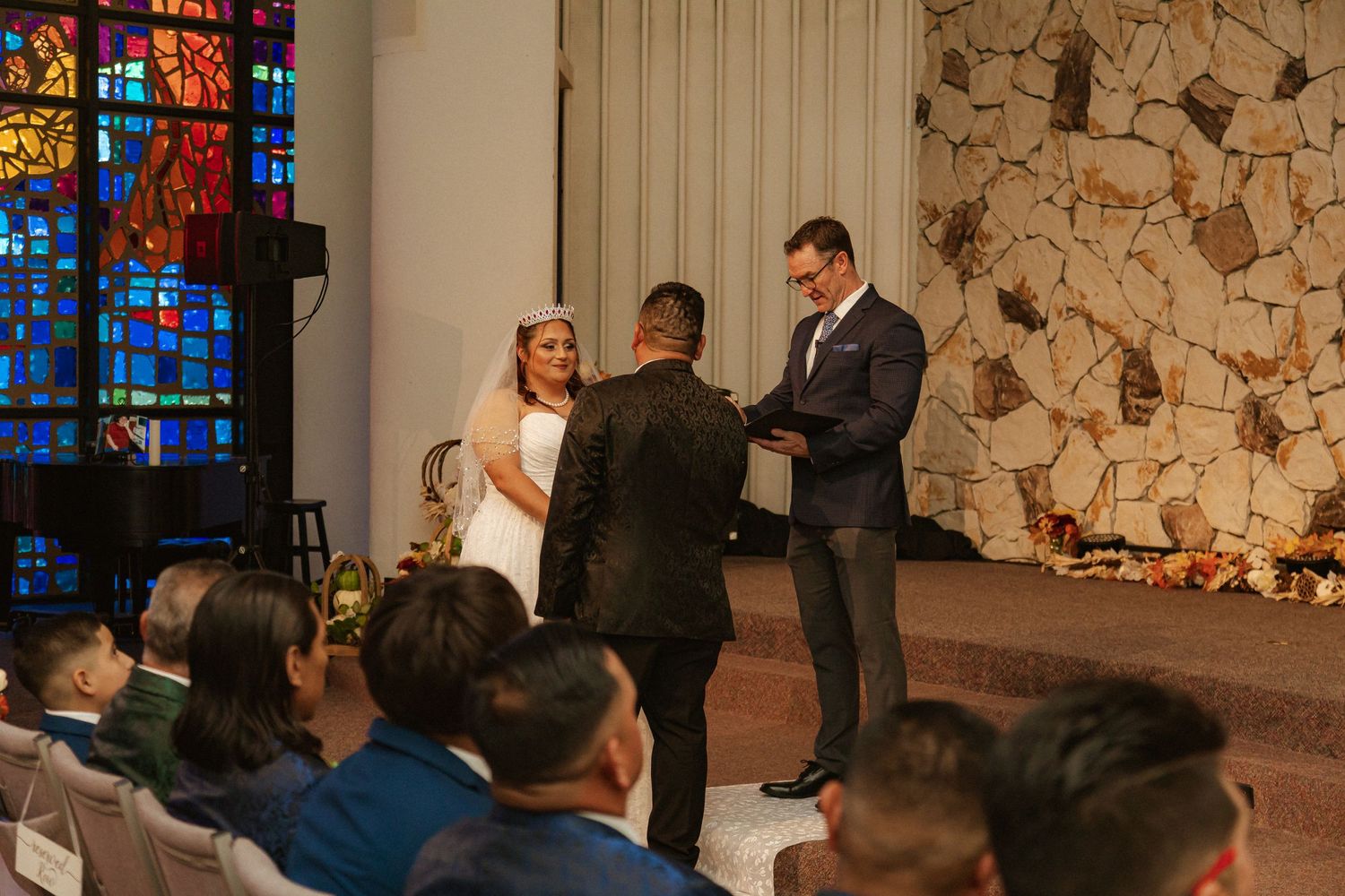 Wedding ceremony conducted by officiant with stone wall and stained glass windows in chapel setting.