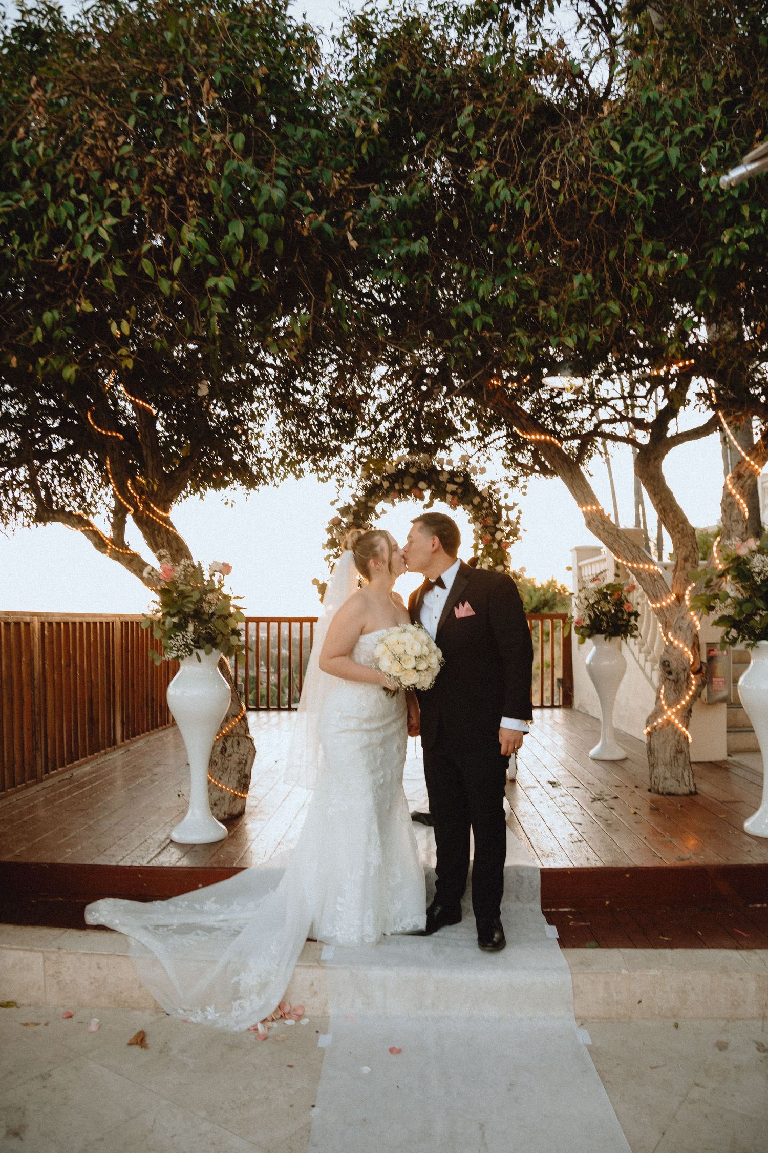 Newlyweds stand together on a terrace with decorative trees and white floral arrangements.