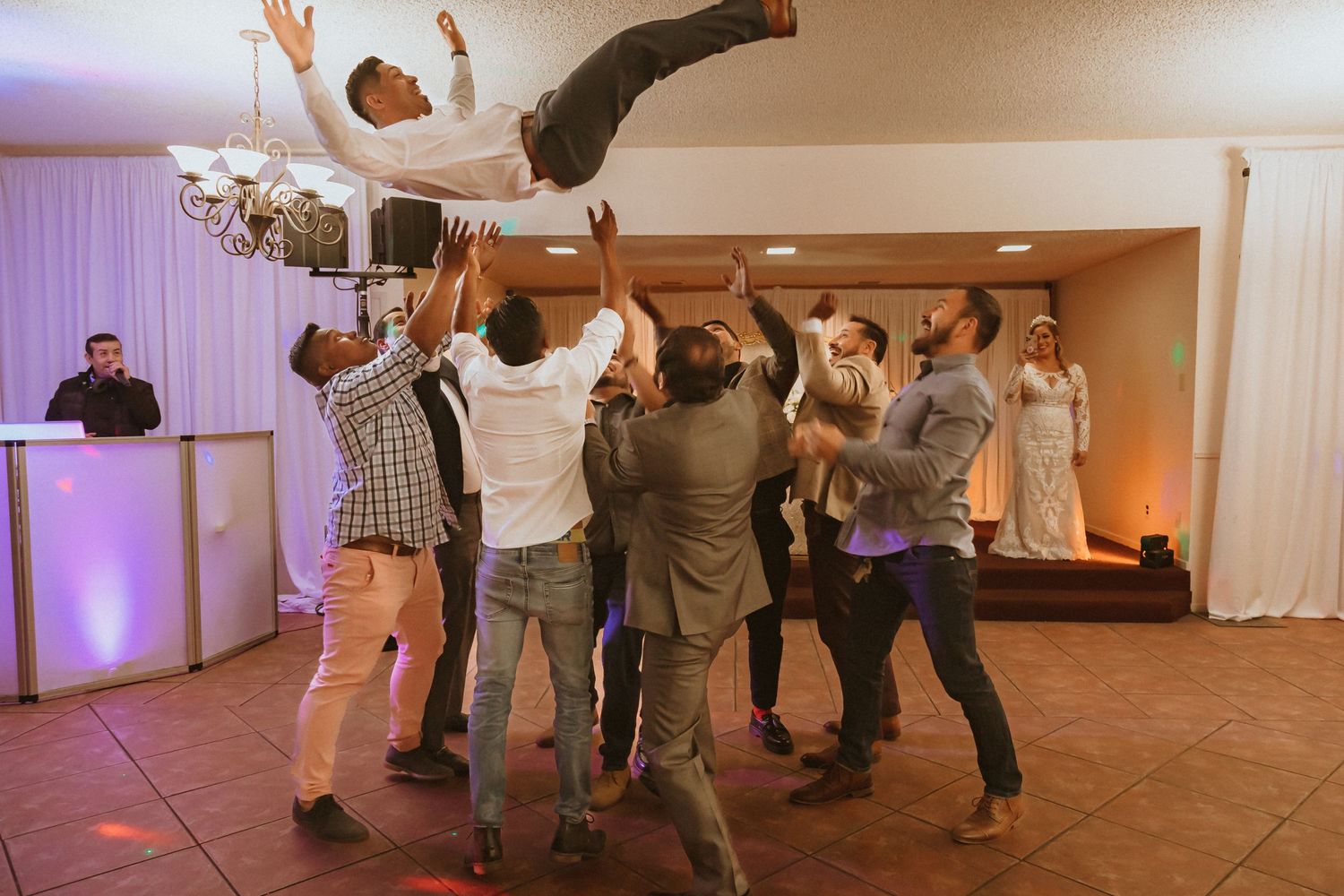 Guests at a celebration toss someone in the air while a DJ booth glows in the background.