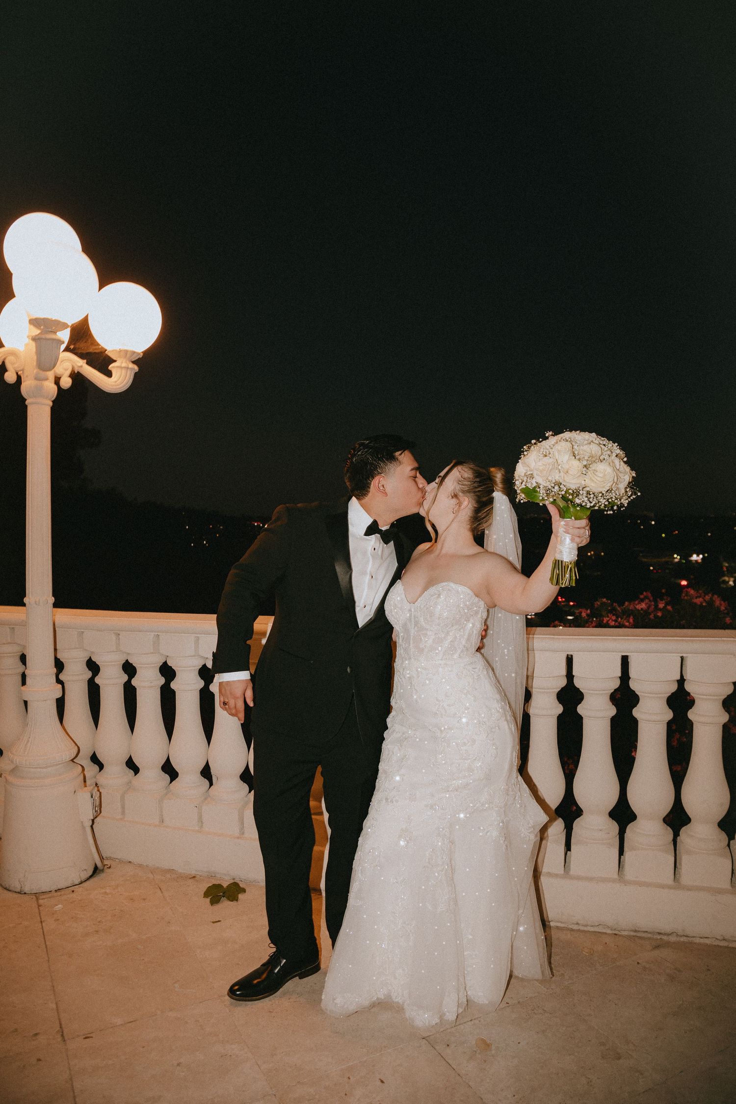 A wedding couple shares a romantic kiss on a balcony with glowing lampposts and city lights behind.