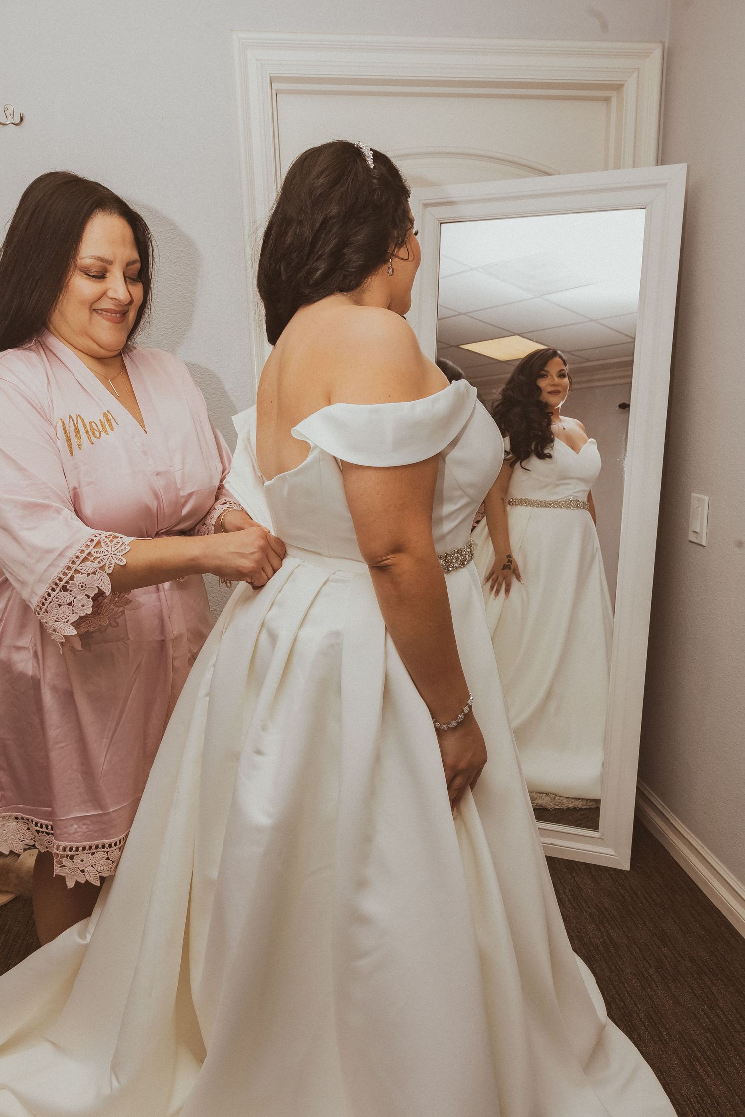 Wedding dress fitting moment captured in mirror reflection with pink robed assistant.