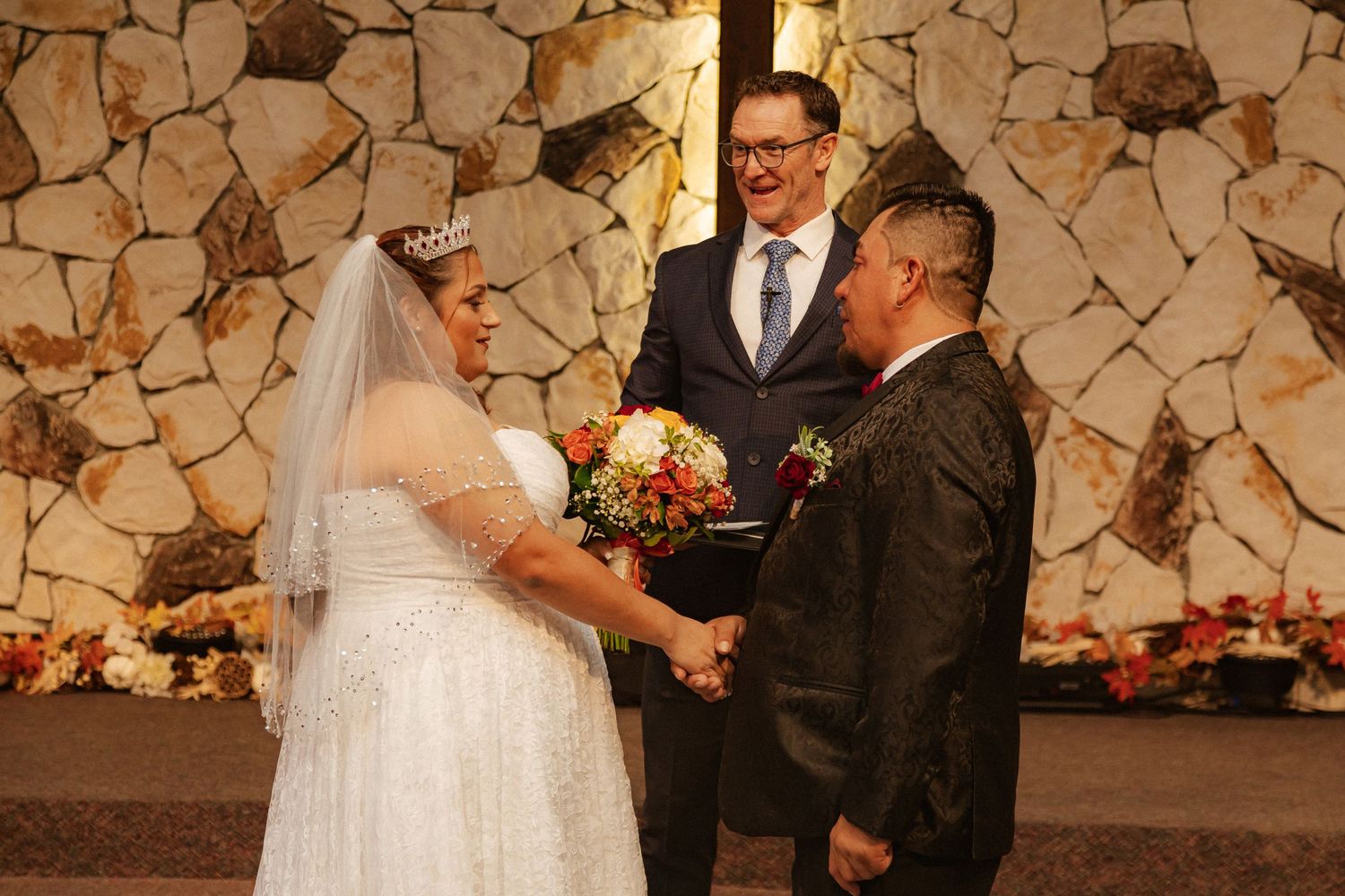 Wedding ceremony in front of stone wall and cross with officiant conducting the service.