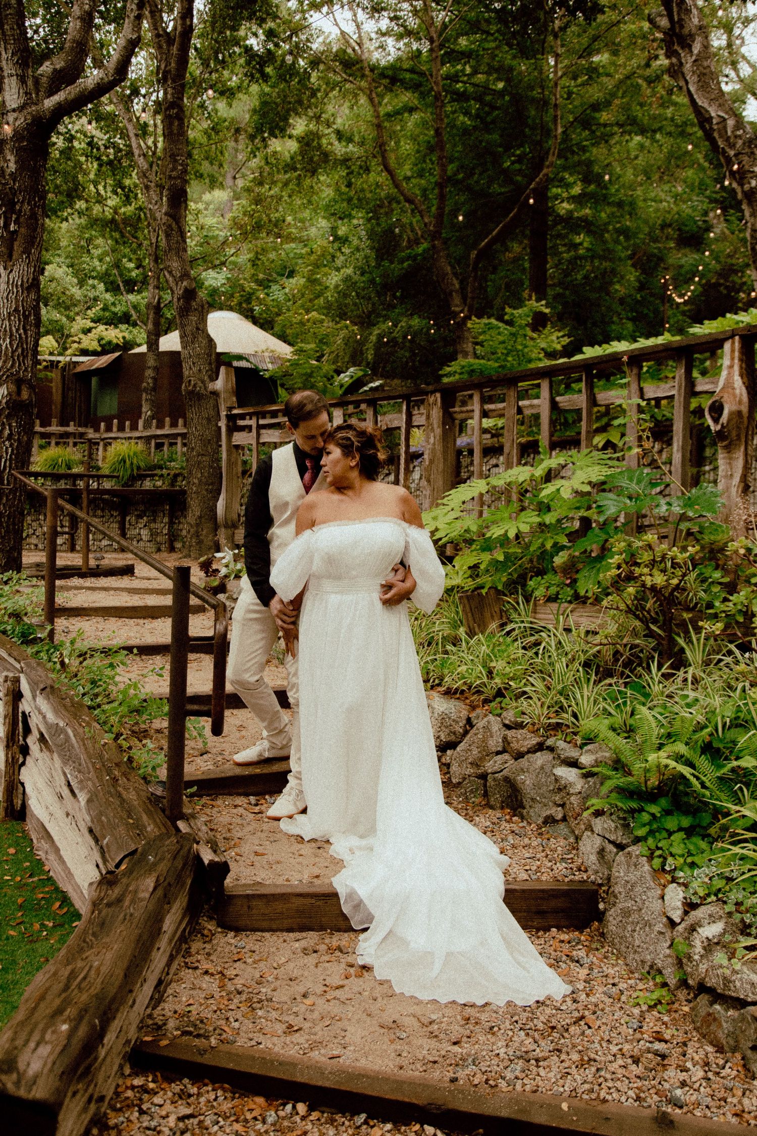 A couple in wedding attire stands on wooden garden steps surrounded by greenery.