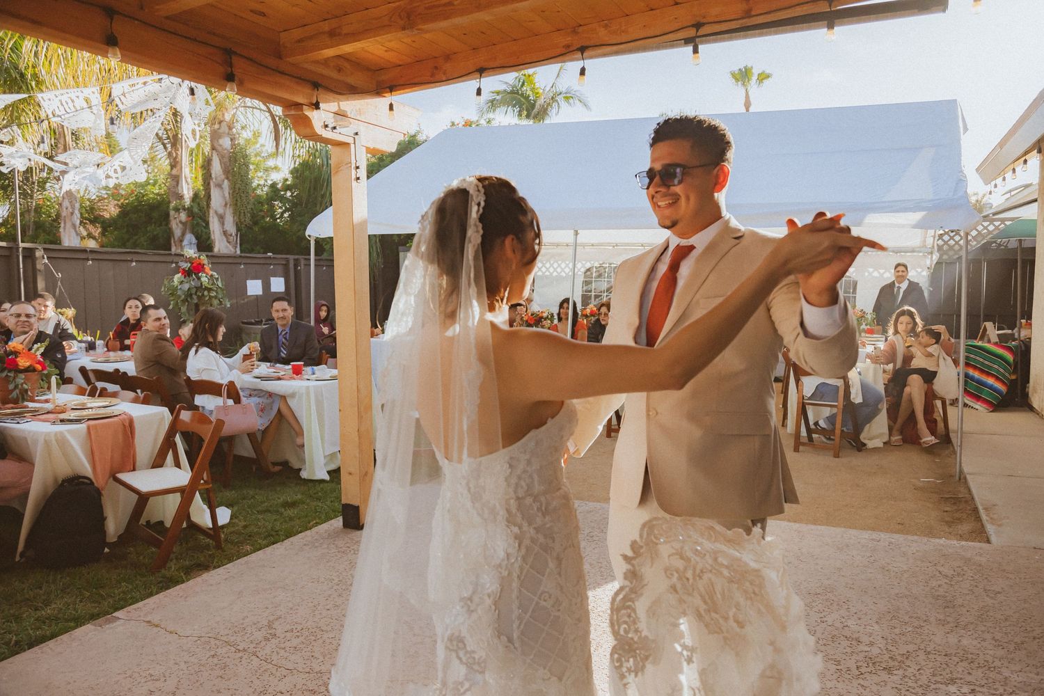 Couple dancing at an outdoor wedding reception under a wooden pergola with guests watching.