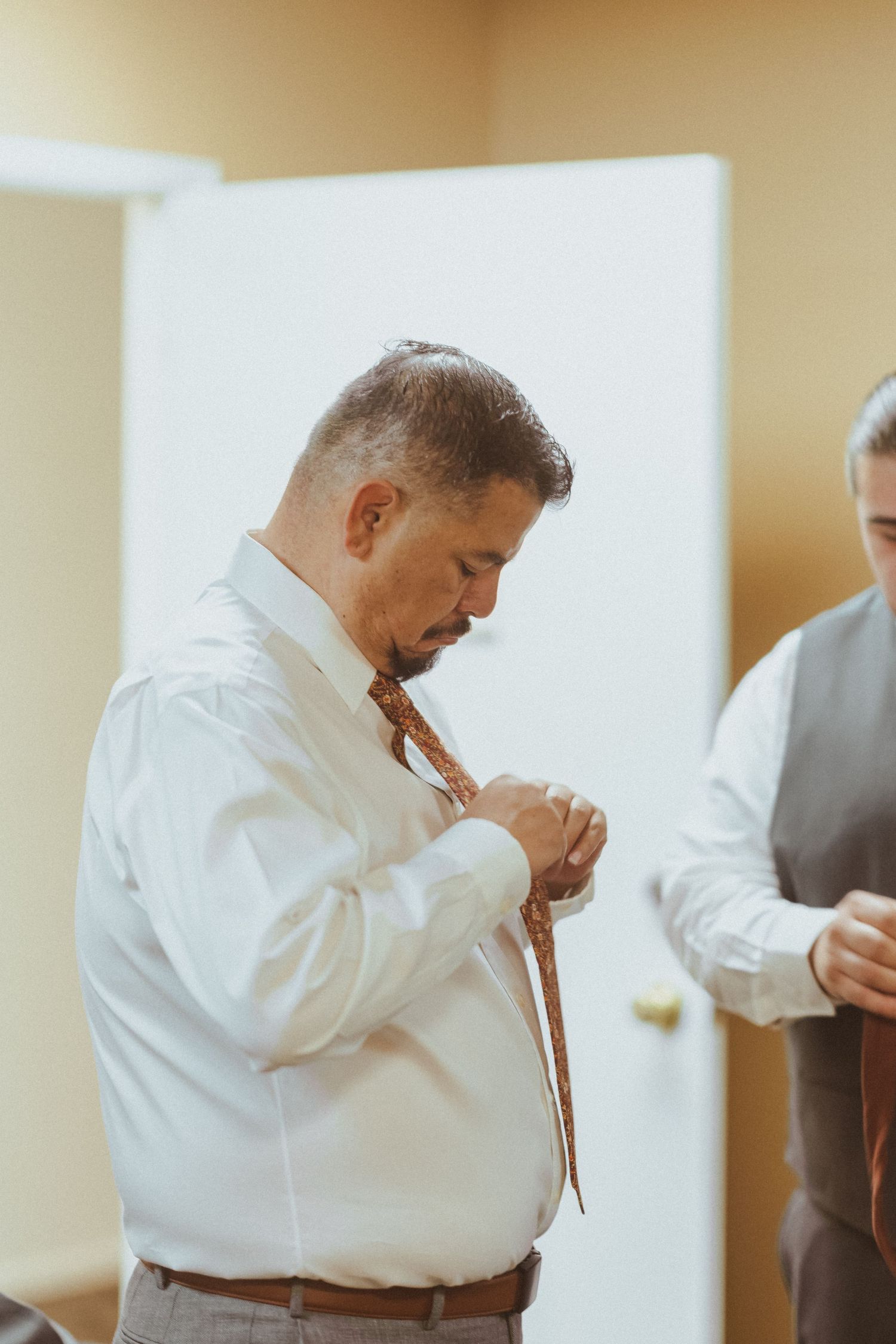 Wedding party member in white dress shirt ties his tie while preparing for formal event.