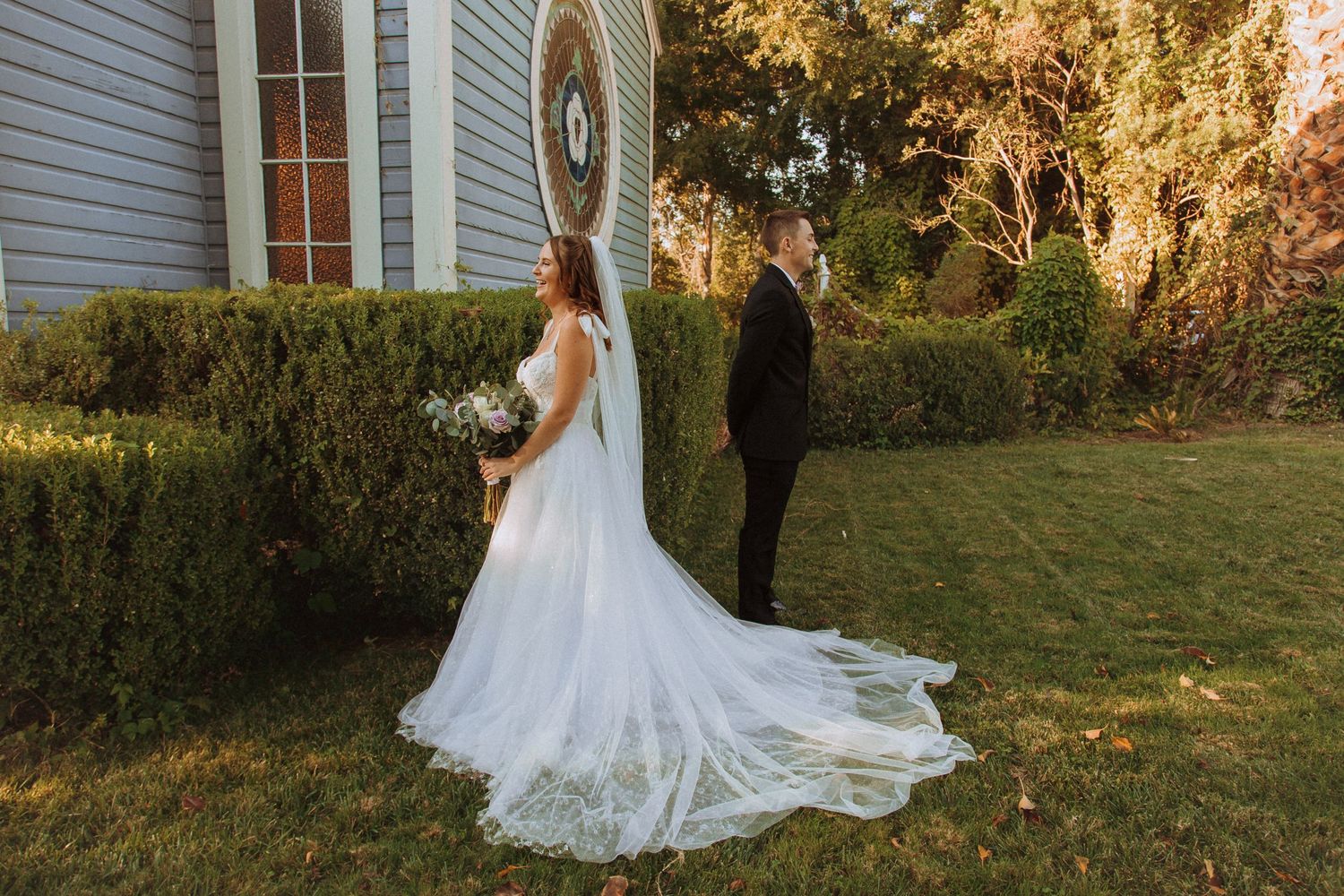 Wedding couple poses separately near chapel showing detailed lace train of bridal gown.