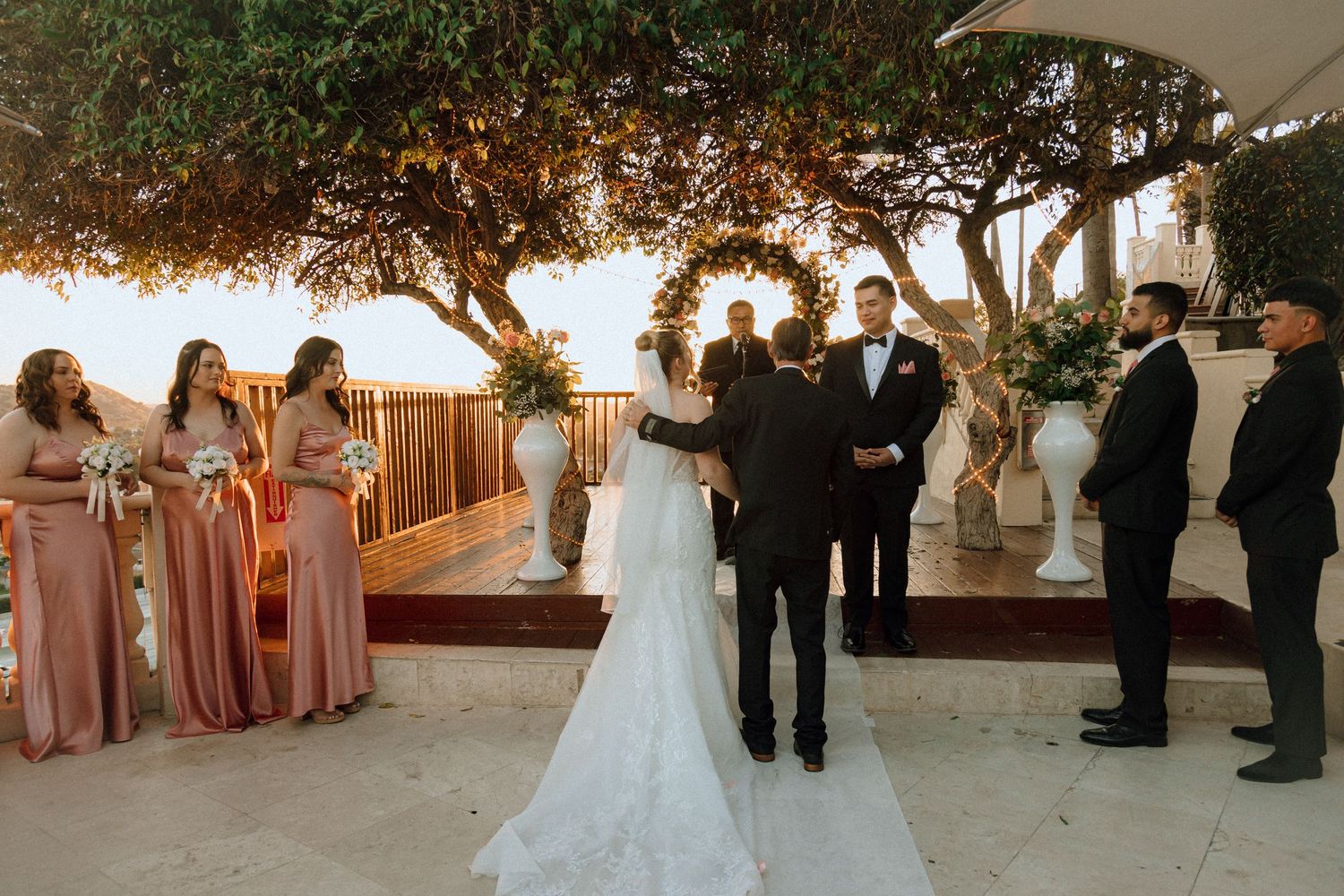 Wedding ceremony taking place at sunset under a tree canopy with guests and wedding party gathered on wooden deck.