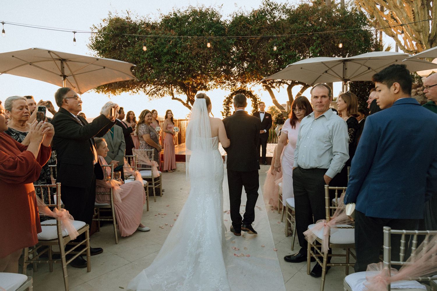 Wedding guests applaud during an outdoor ceremony with string lights and umbrellas overhead.