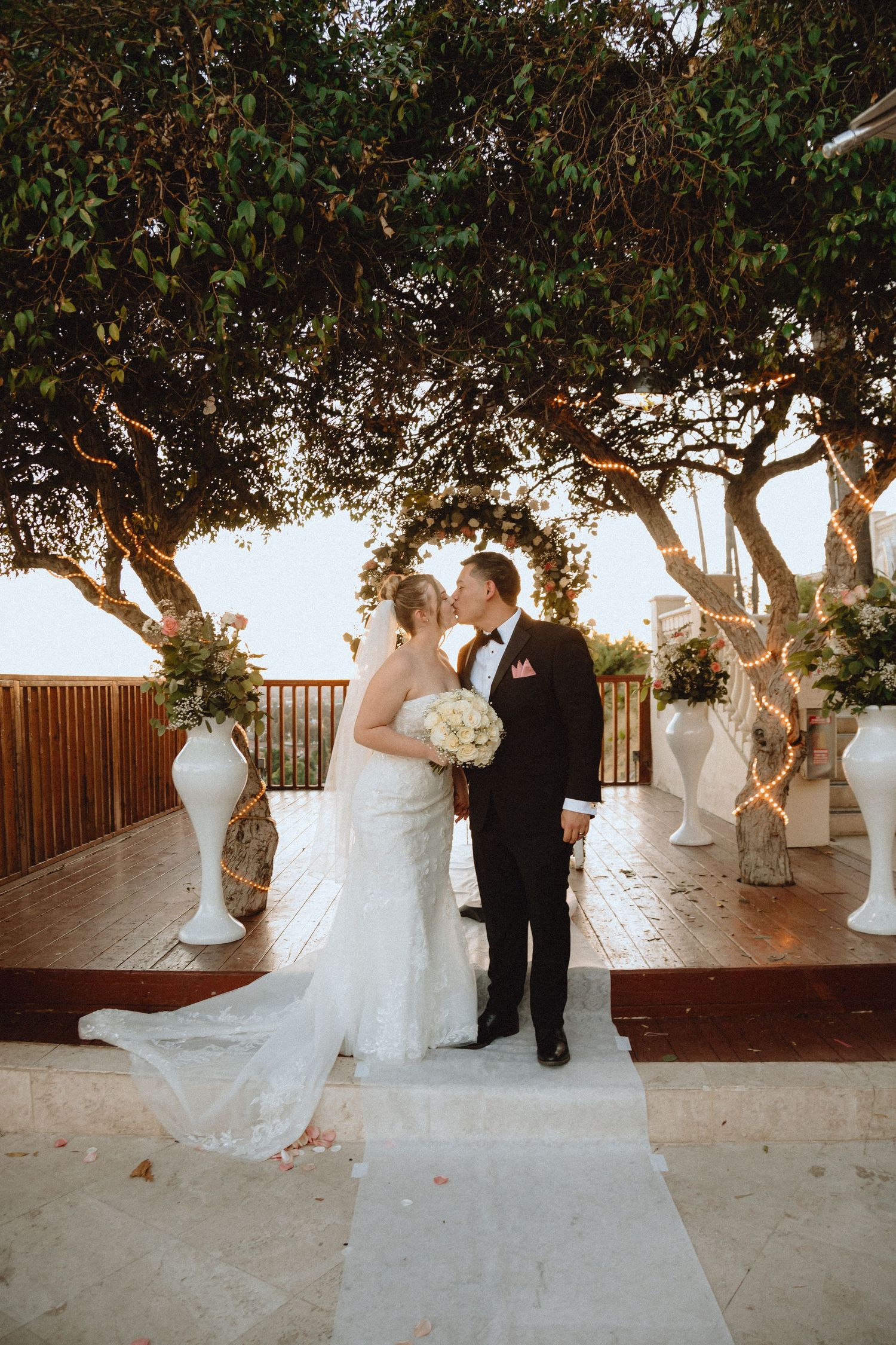 A couple shares a romantic moment under trees at their outdoor wedding ceremony with decorative white vases.