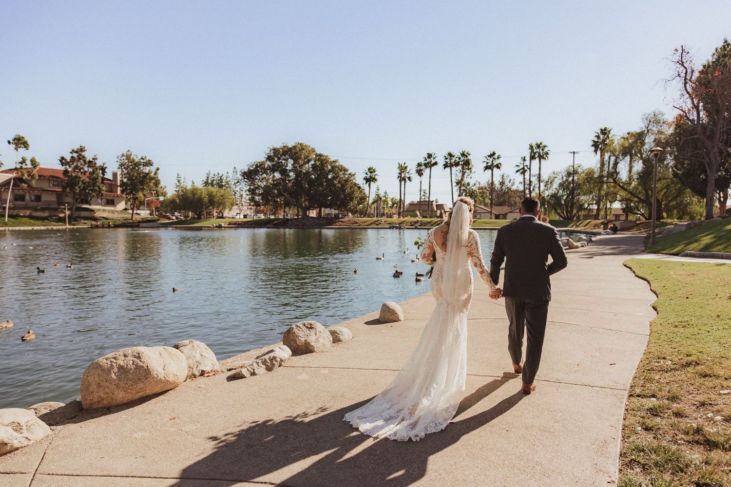 Newlyweds walking together on a scenic pathway beside a tranquil lake with palm trees.