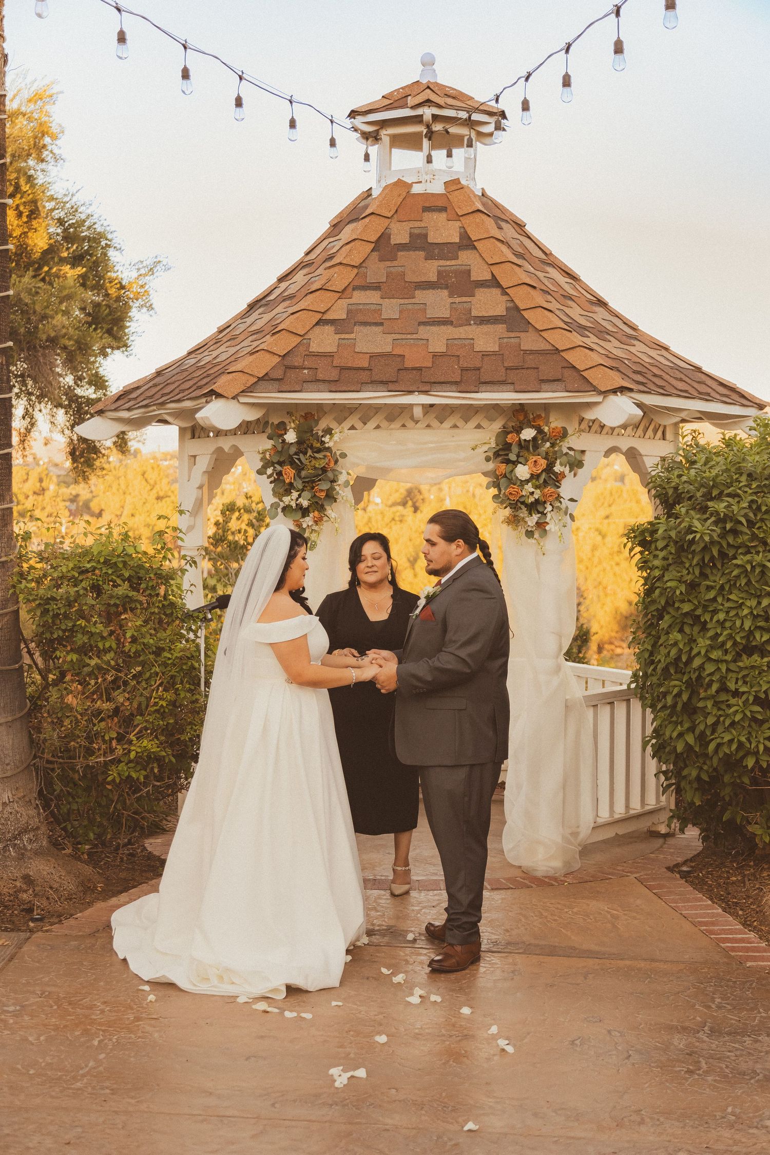 Intimate wedding ceremony moment under an octagonal gazebo with string lights at sunset.