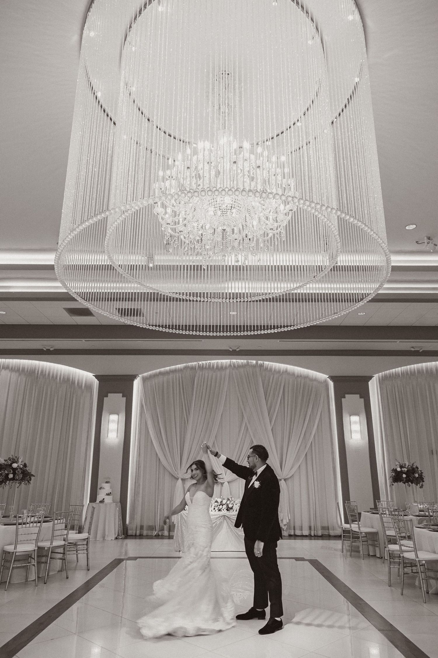 Black and white wedding photo showcasing elegant ballroom with crystal chandelier and draped walls.
