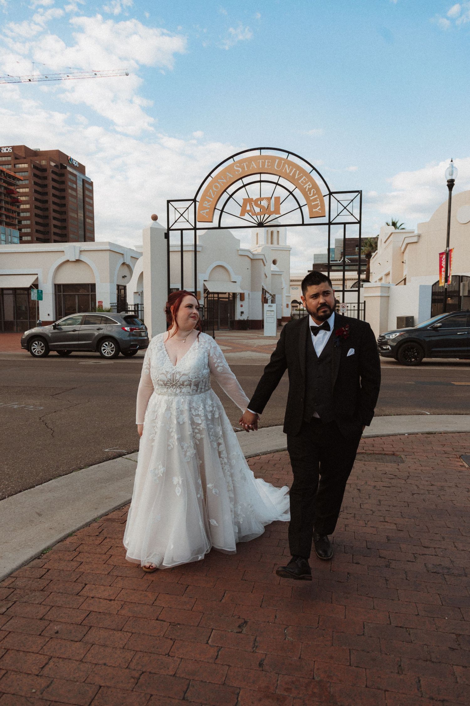 Newlyweds walk hand in hand on brick pavement with an art deco archway in the background.