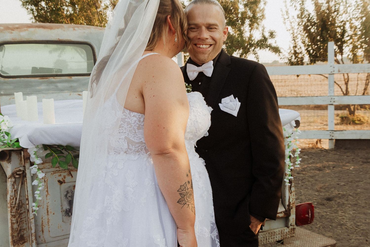 A bride in a white gown and groom in black tuxedo share a joyful moment during their countryside wedding.