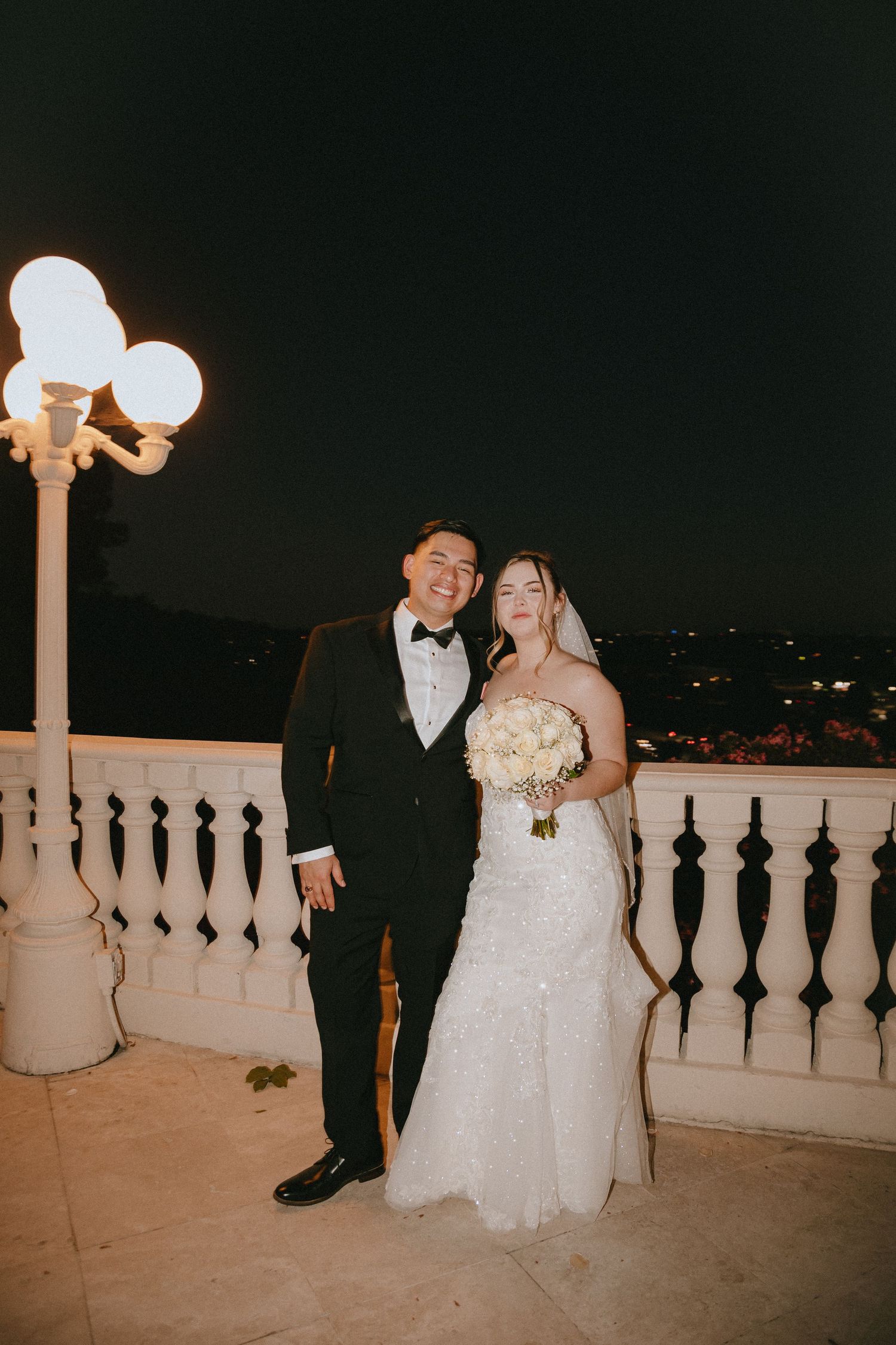 Newlyweds pose on a balcony at night under glowing vintage streetlights.