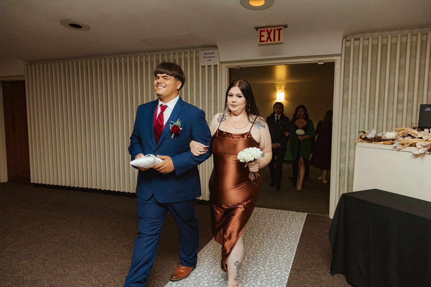 Wedding party members process down decorated hallway in formal evening wear.