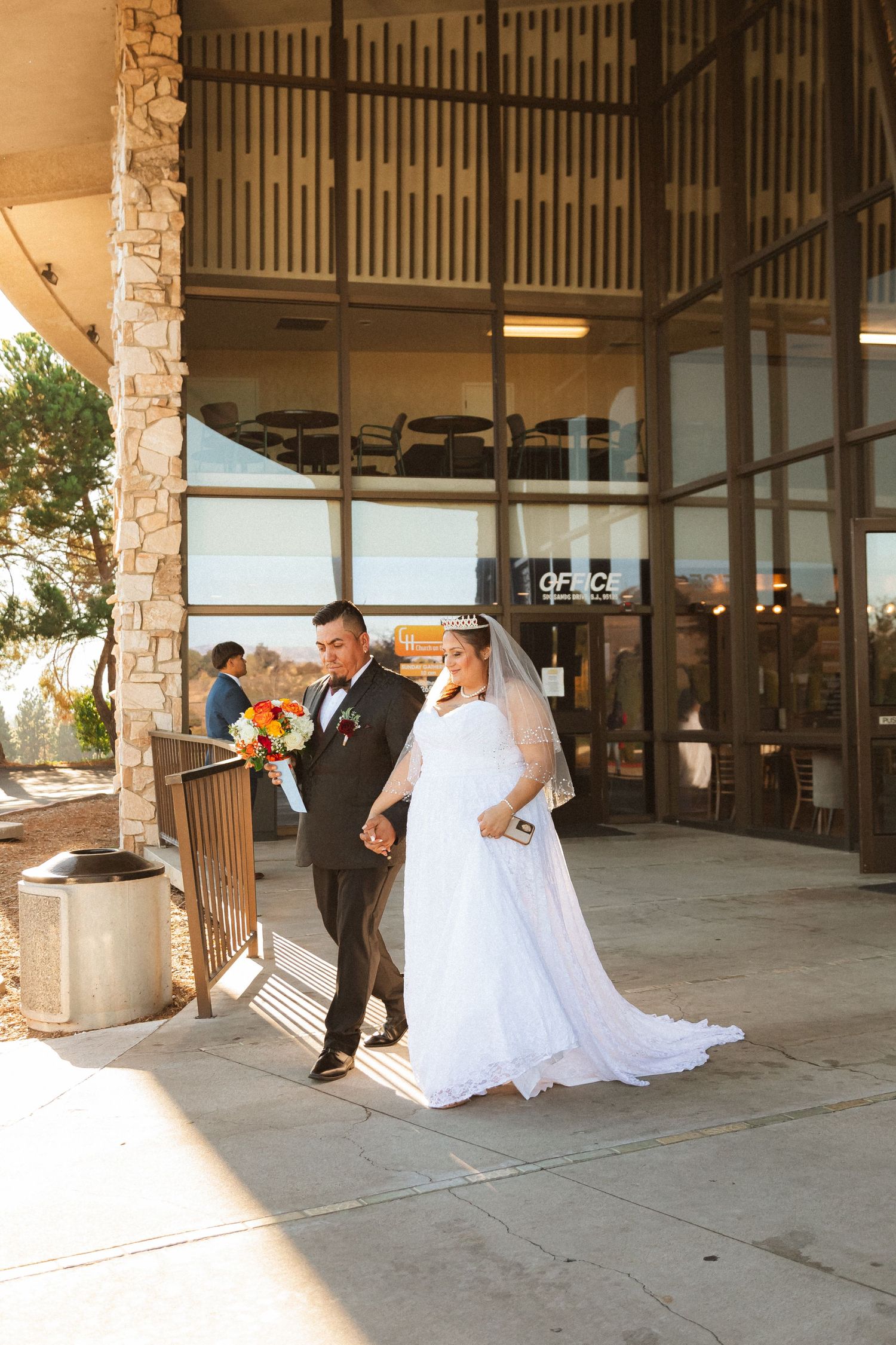A couple in wedding attire walk together on a covered patio area with stone columns in warm sunlight.