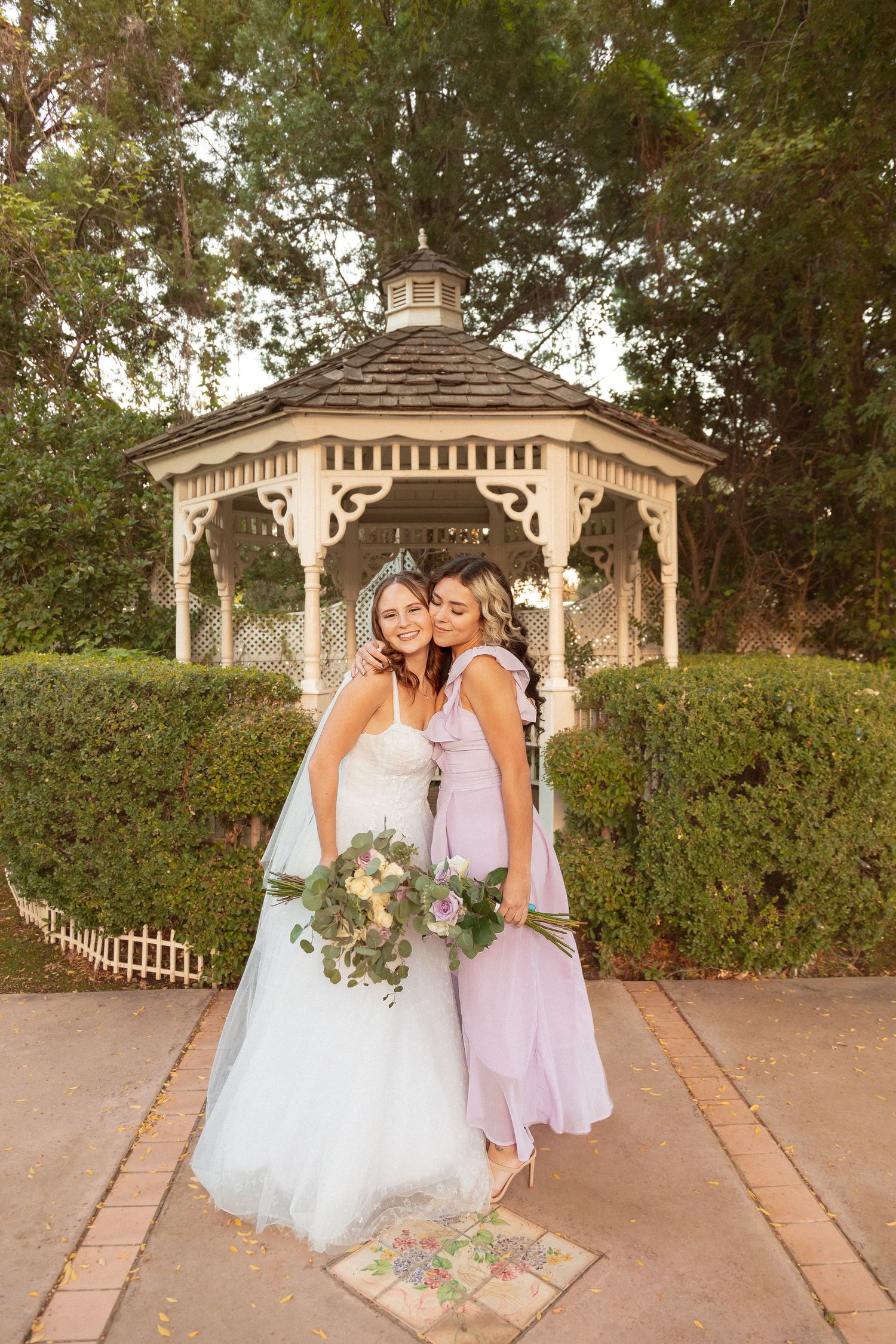 Two people in formal wear embrace near a garden gazebo surrounded by hedges.