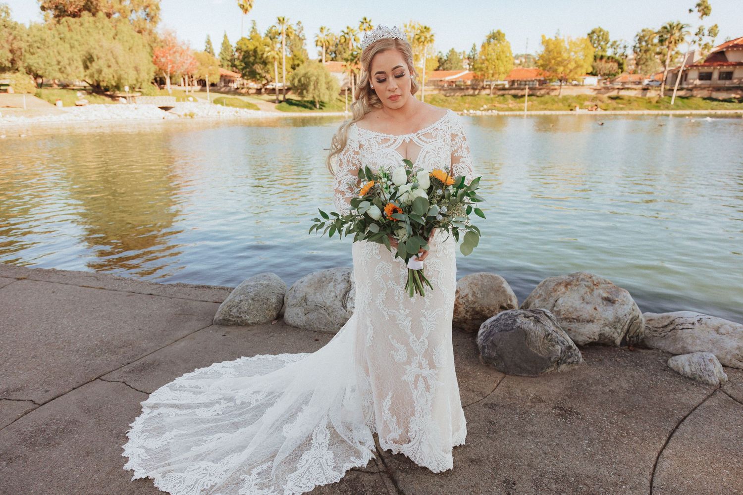Lakeside bridal portrait showcasing a fitted lace gown and mixed floral bouquet against fall foliage.