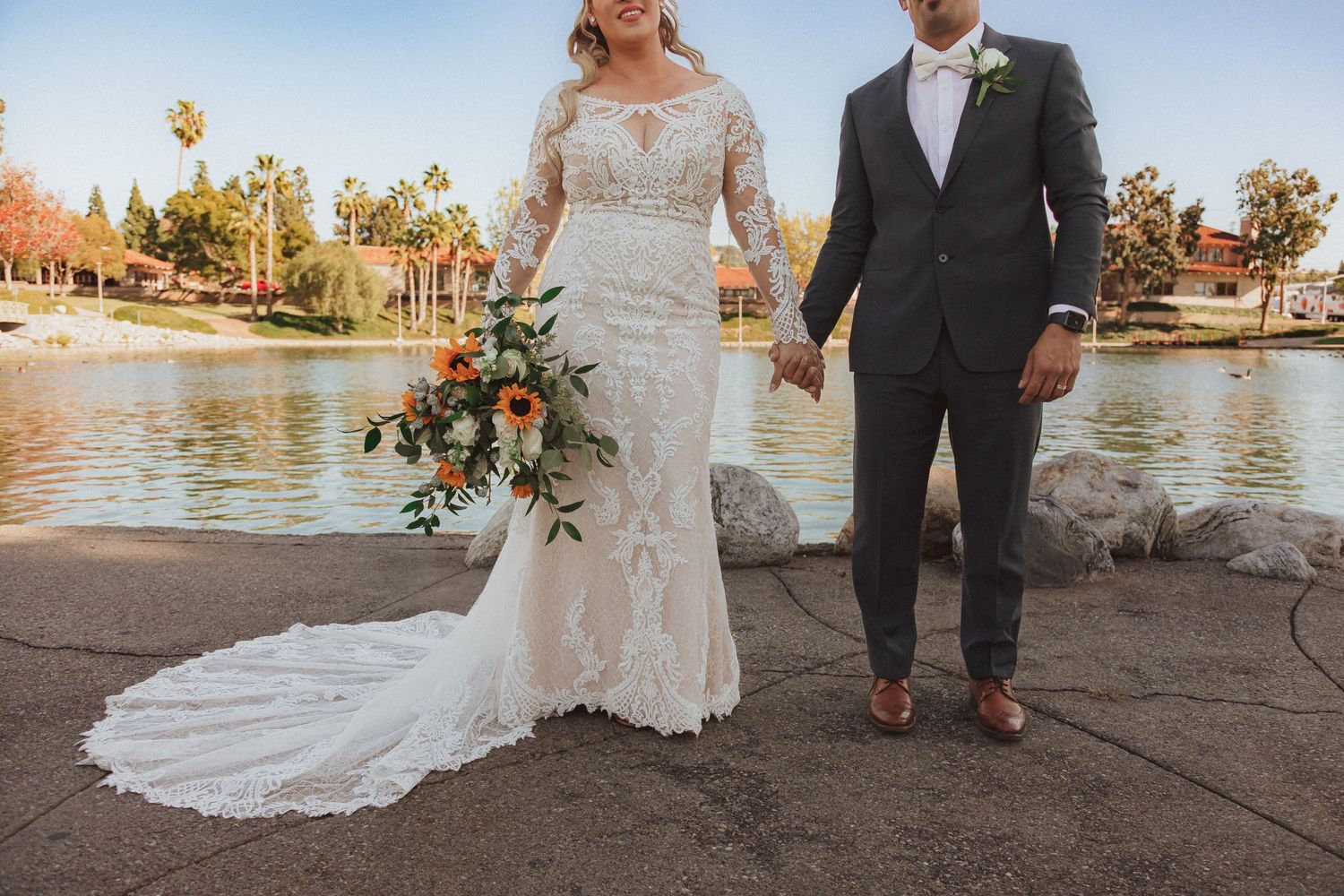 Full-length shot of wedding couple by lake showing bride's detailed lace gown with long train.