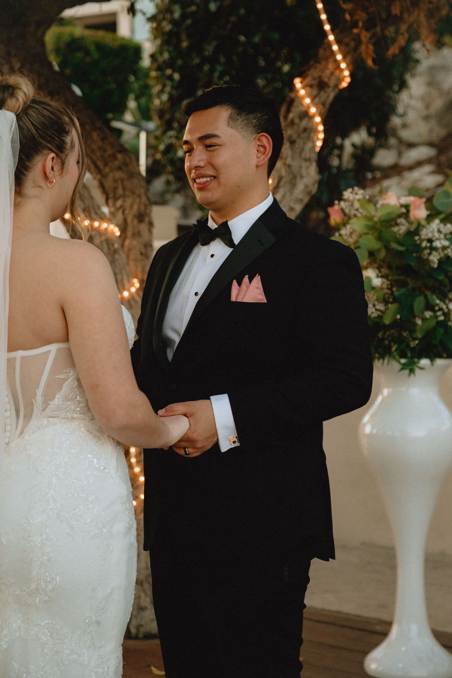 A romantic wedding moment as the couple holds hands during their ceremony under twinkling string lights.