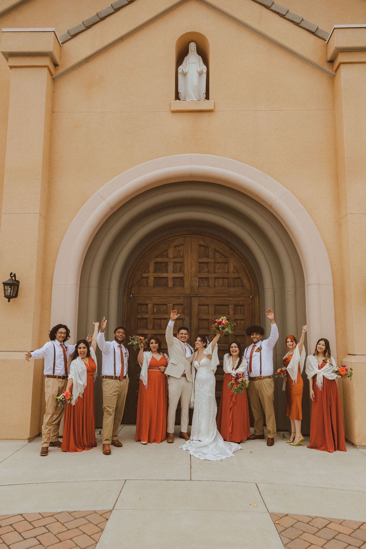 Joyful wedding party photo with coordinated coral and khaki outfits against church facade.