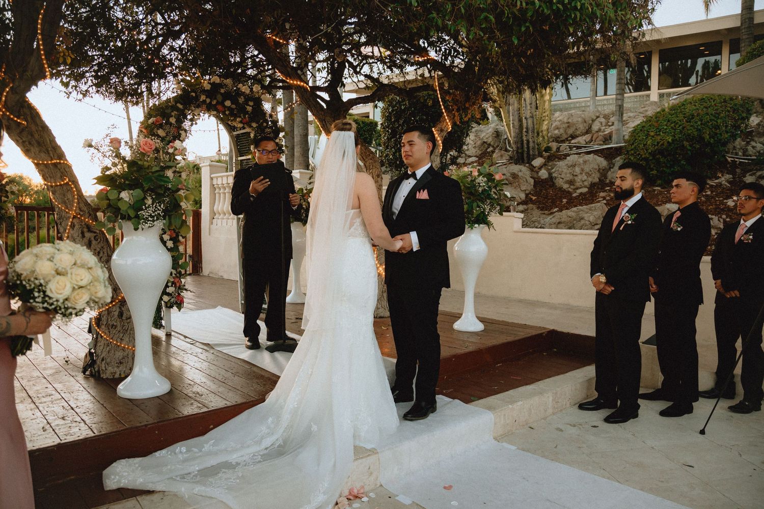 Outdoor wedding ceremony with decorative white pillars and string lights among trees.