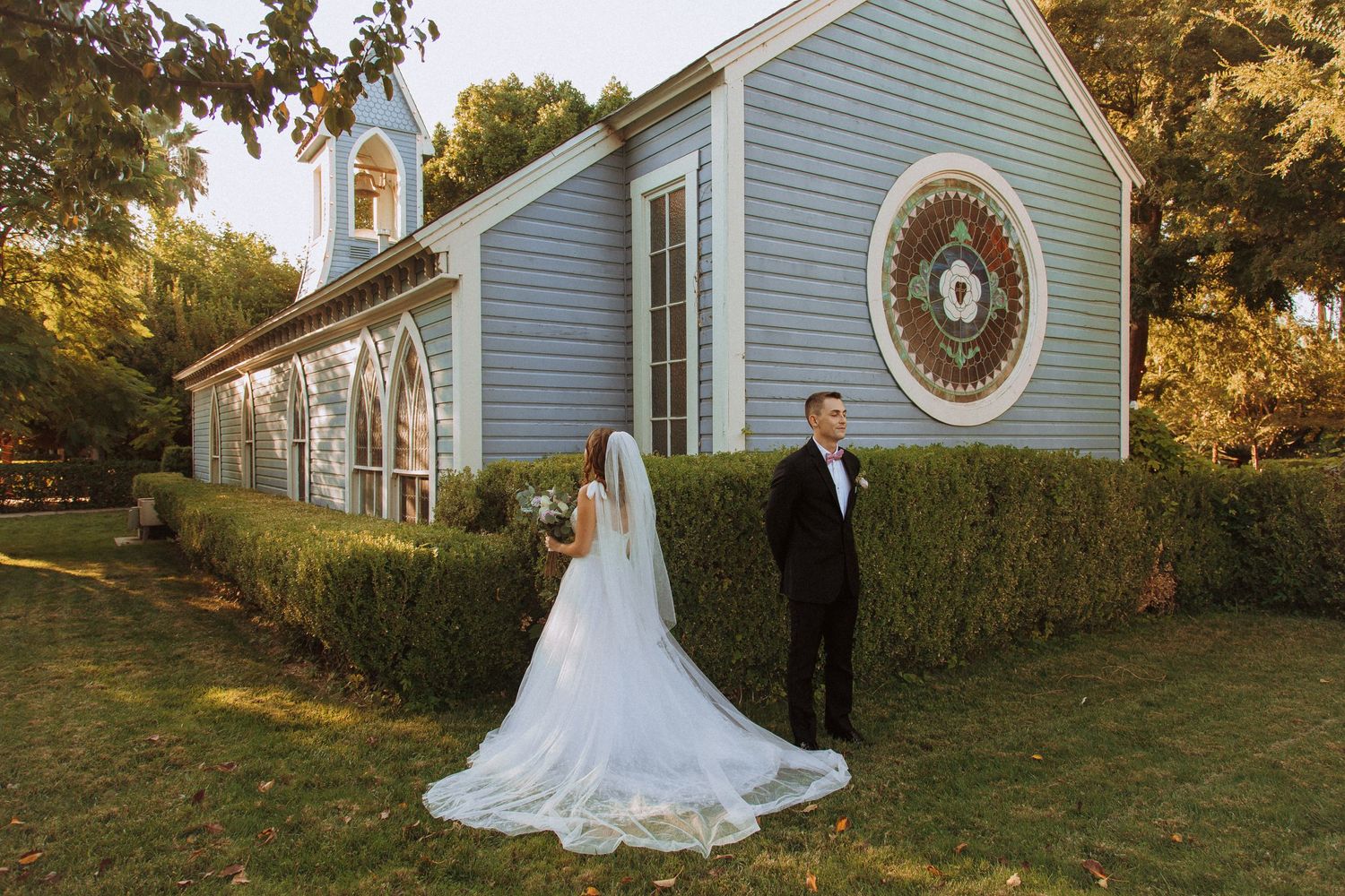 Couple poses in formal wedding attire outside a light blue chapel with round window.