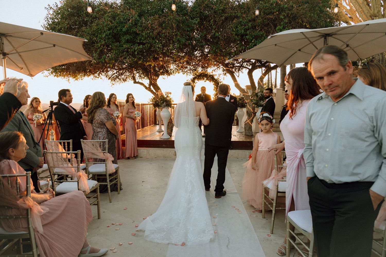 An outdoor wedding ceremony procession with guests lining a white aisle runner.