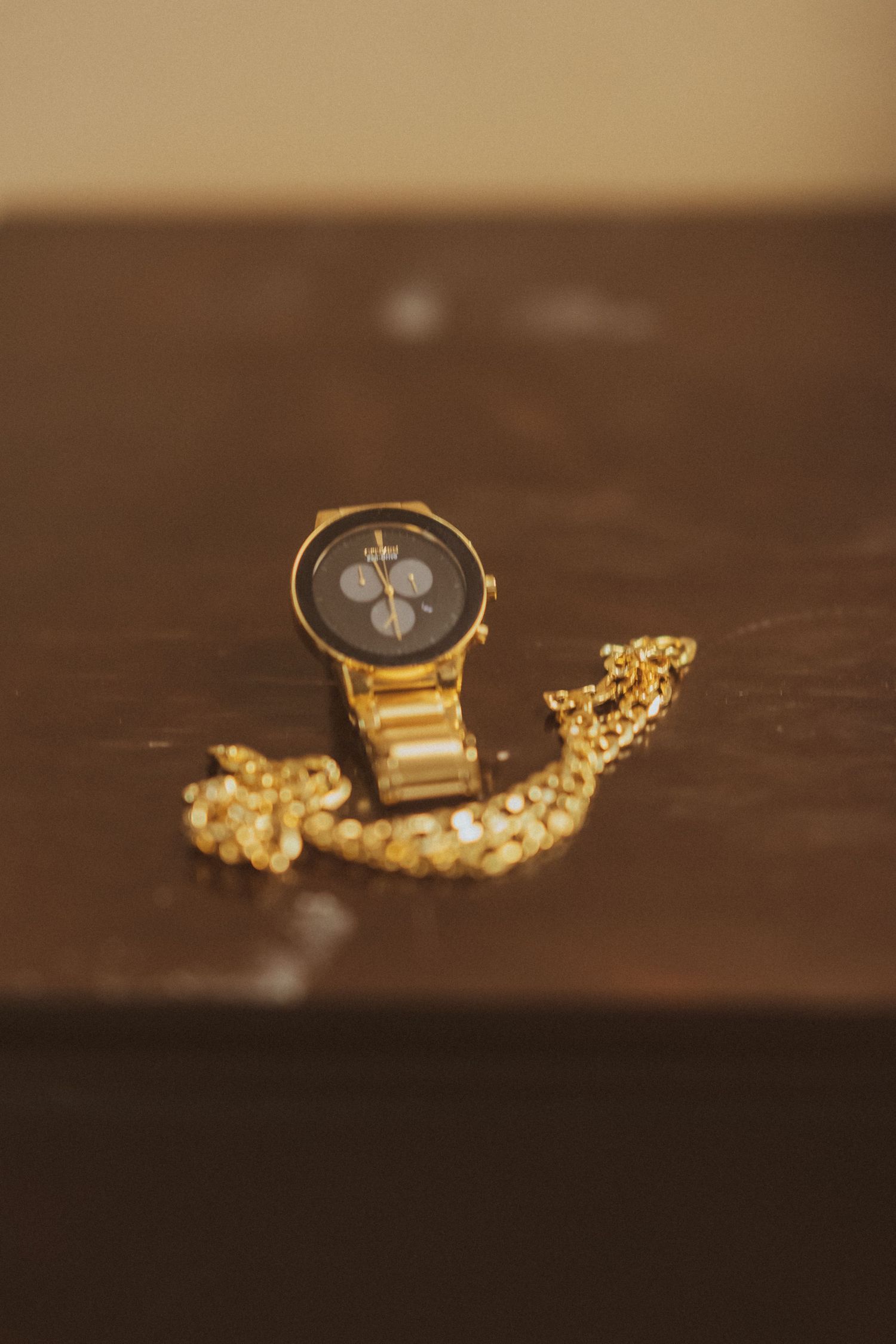 Gold watch with black face and delicate chain necklace on dark surface.