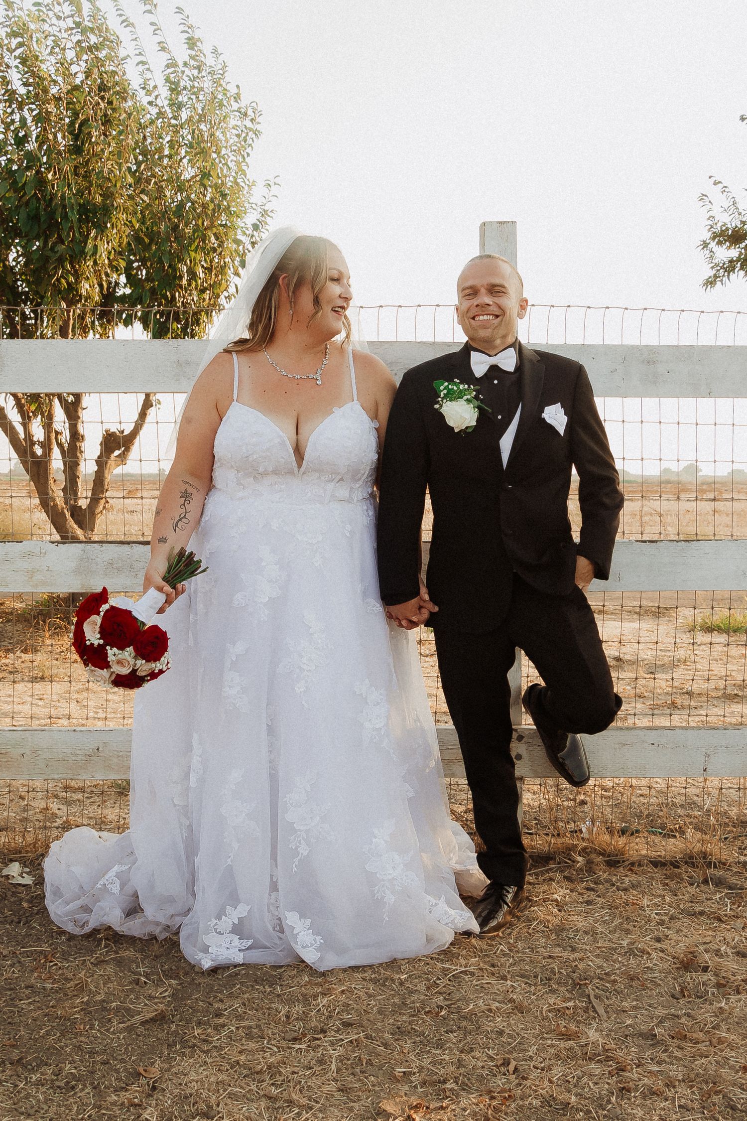 A couple poses for wedding photos by a white fence, with the bride holding a red bouquet.