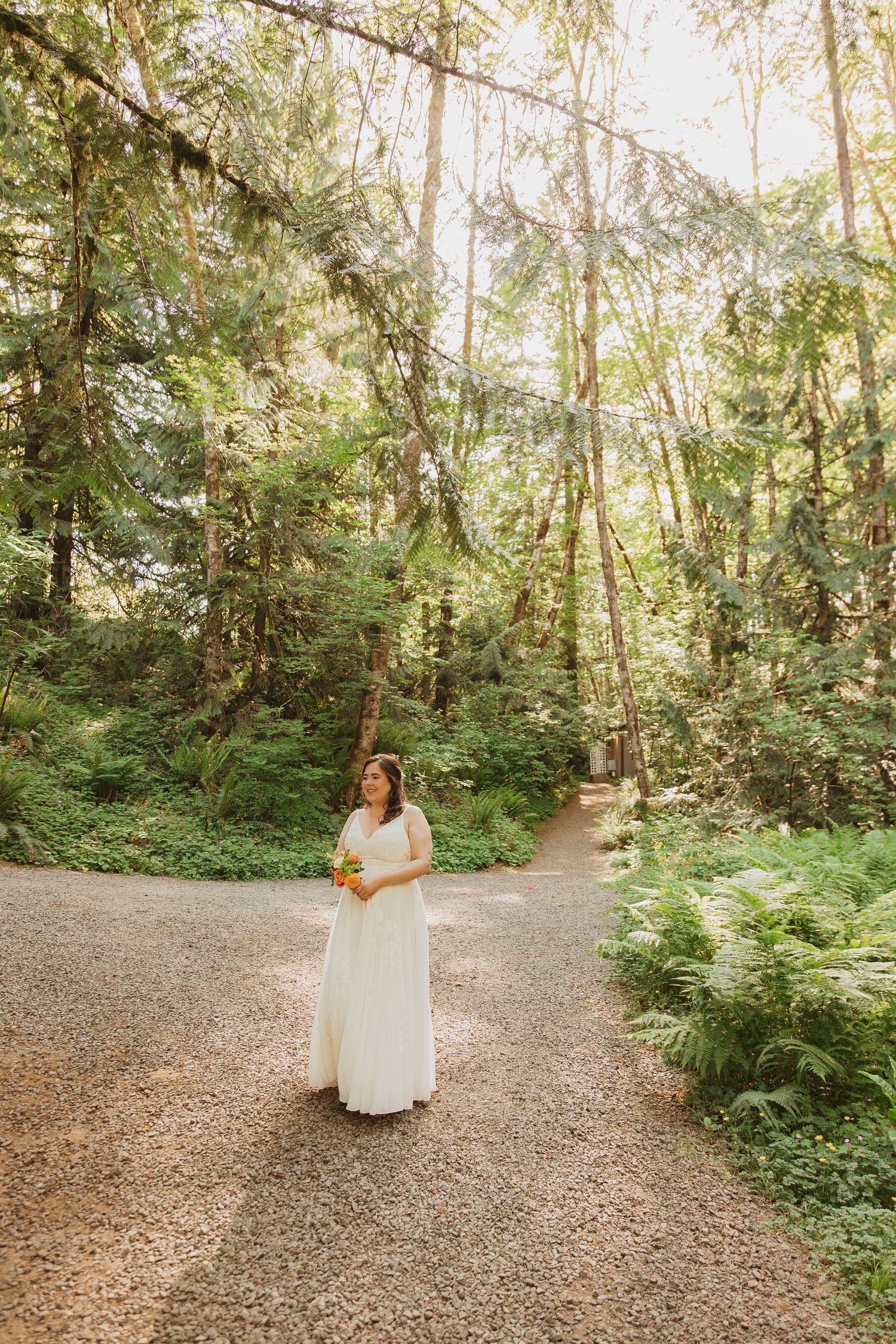Bride holding yellow bouquet stands on forest path surrounded by tall evergreen trees.